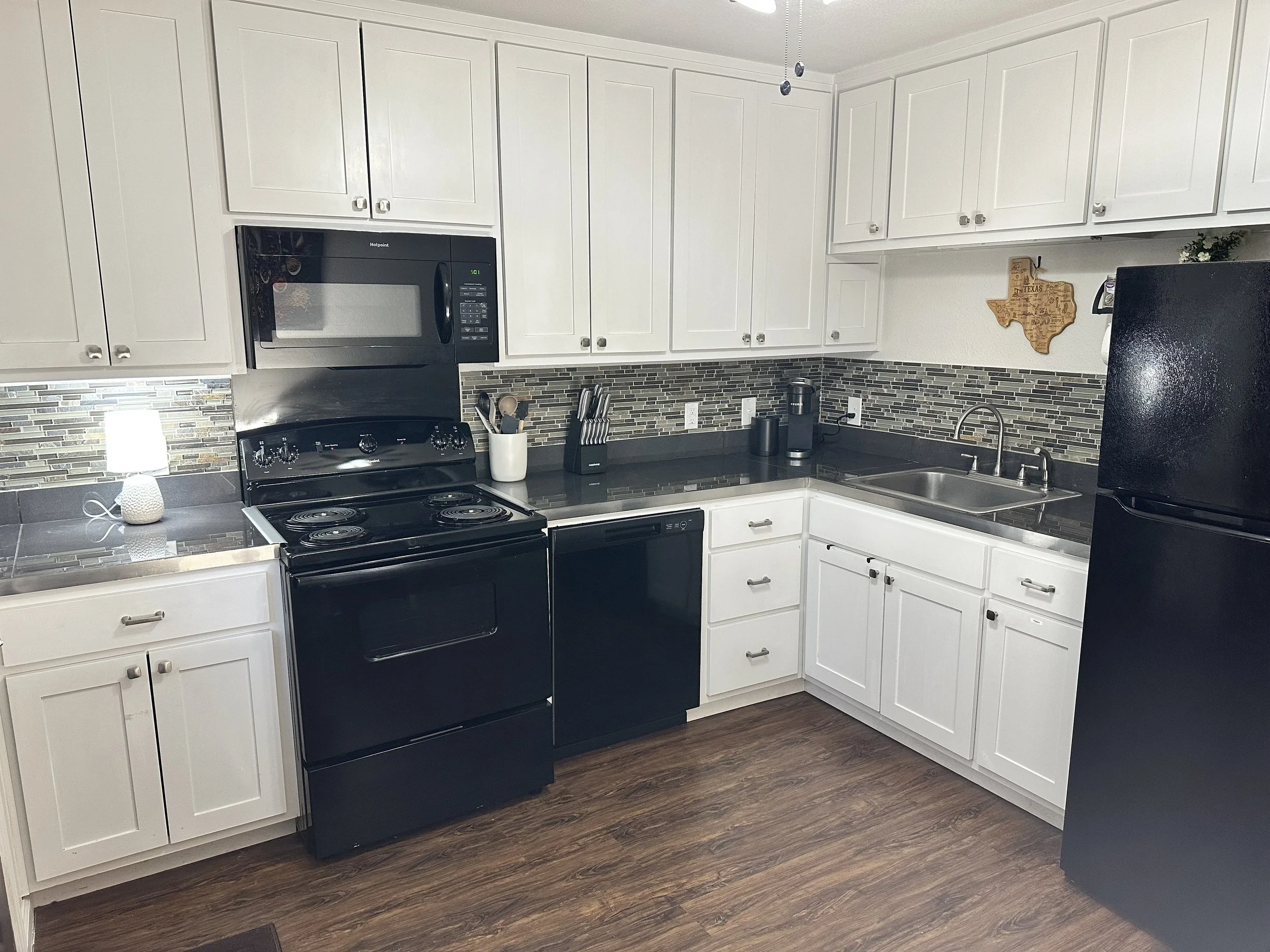 Kitchen with white cabinets, black stove, microwave, black refrigerator, tiled backsplash, stainless steel sink, and wooden flooring.