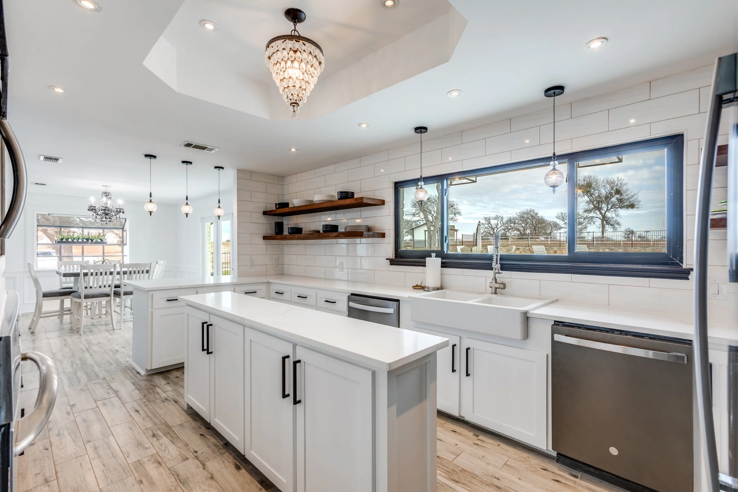 Modern kitchen with white cabinets, a large island, pendant lights, and a window showing an outdoor view with trees and a fence.