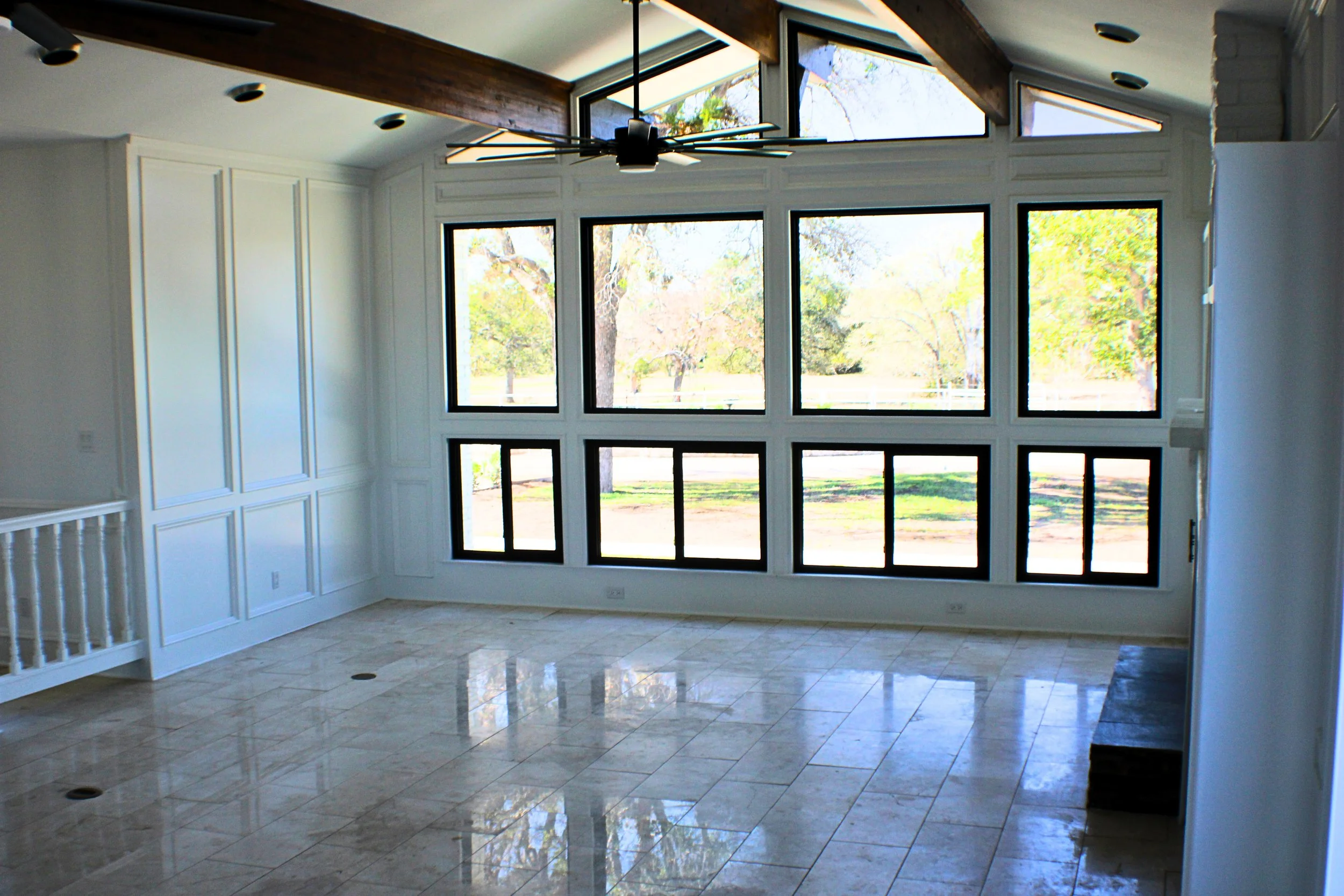 Interior of a room with large windows showcasing a view of trees and a field outside, a modern ceiling fan, and tiled floors.