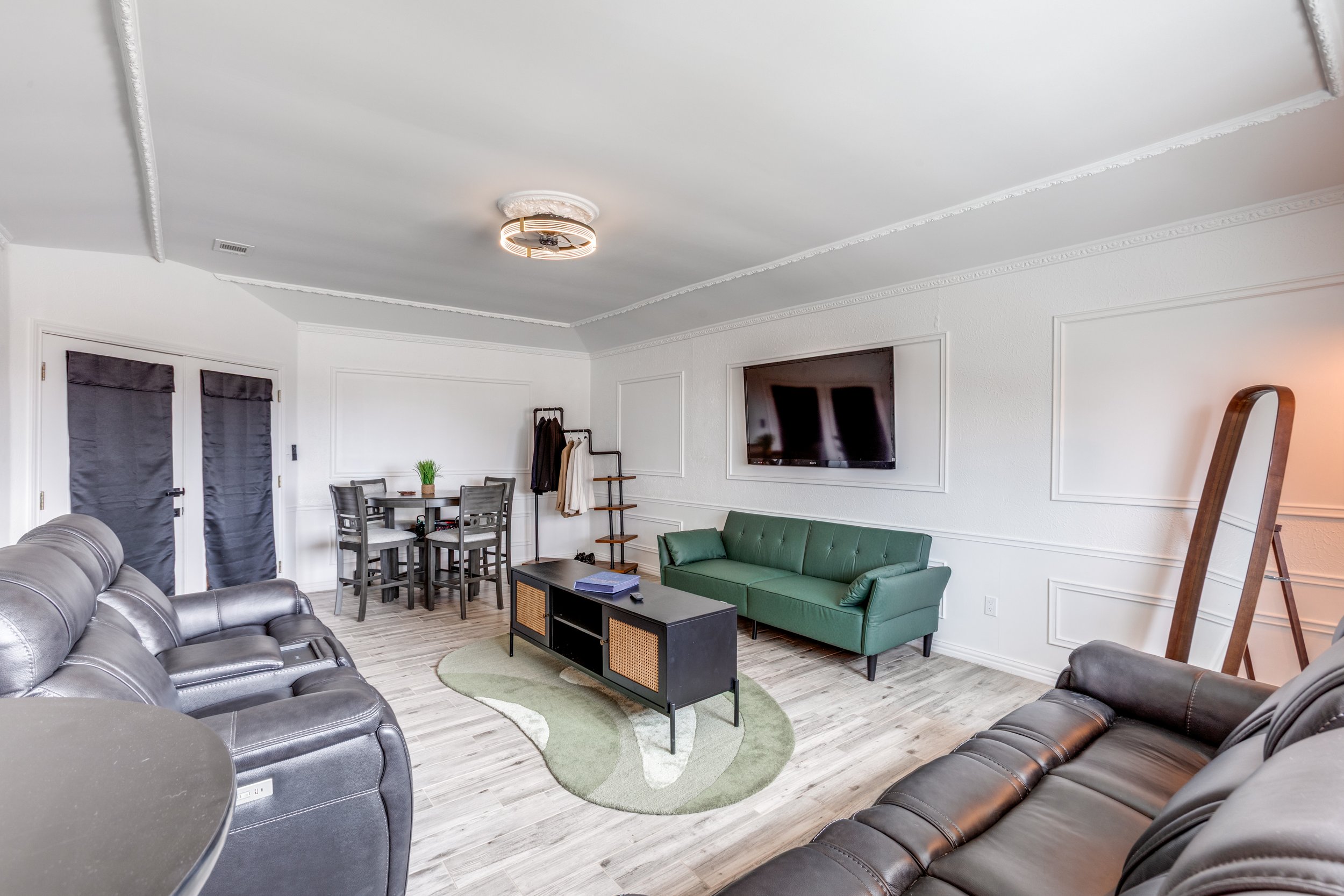 A living room with leather sofas, a green sofa, a wall-mounted TV, a small round table, a rug, and a hallway with doors covered with dark curtains. There is a ceiling light and a coat rack near the dining table.