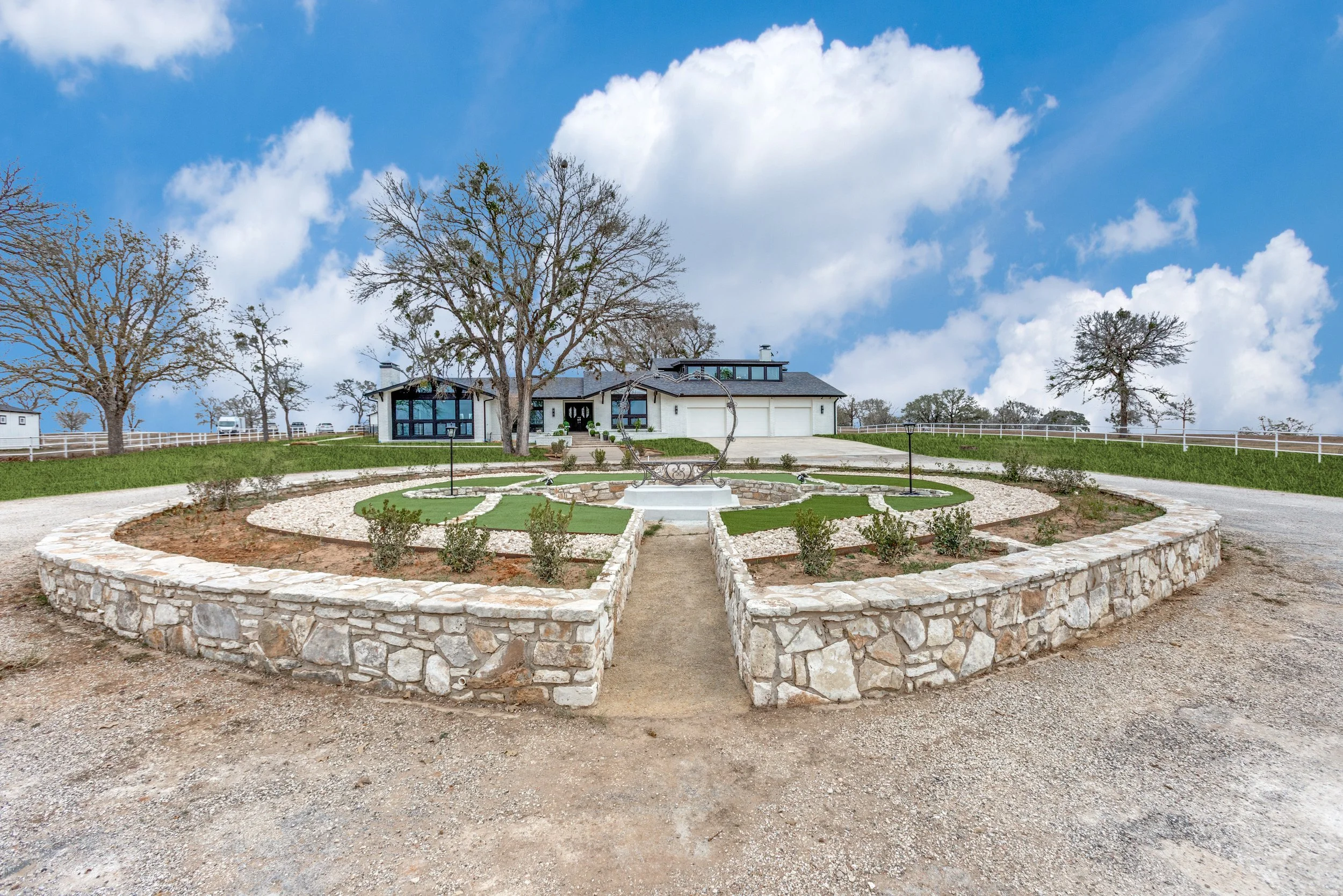 A large, modern two-story house with a dark roof and white exterior walls. The house is surrounded by a landscaped yard with bare trees, a garden bed with small shrubs, and a stone pathway. The sky above is bright blue with scattered white clouds.