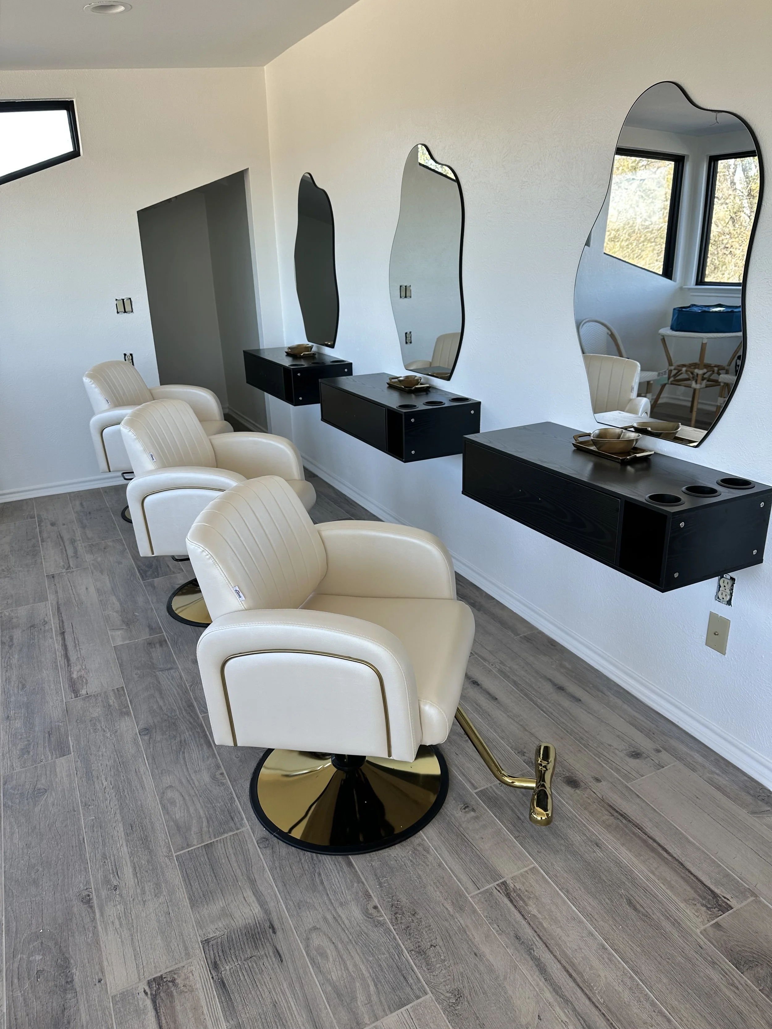 A salon interior with four white chairs with gold accents facing a wall with three black floating station mirrors and small counters. The room has wood-look tile flooring and windows allowing natural light.