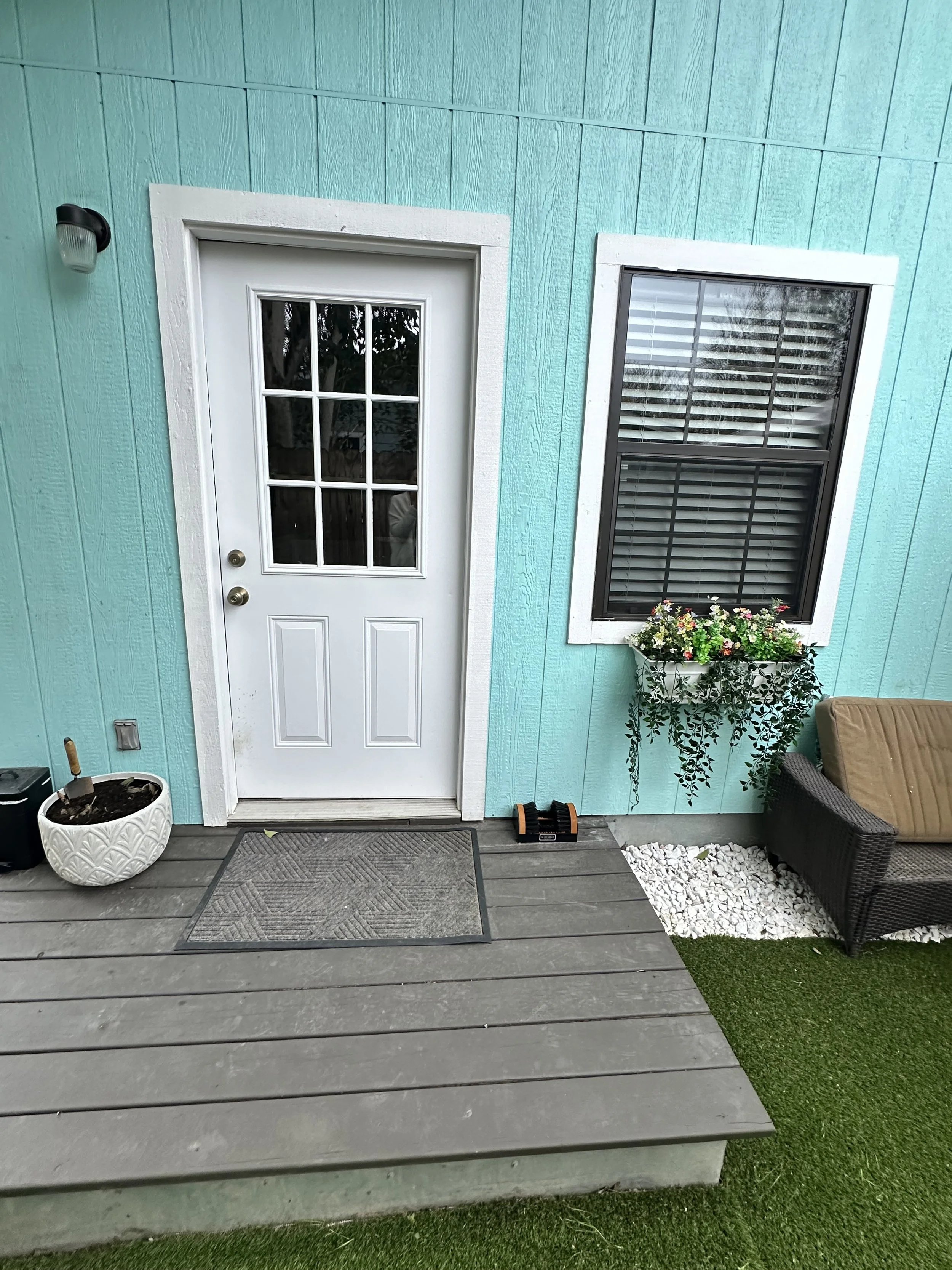 Exterior view of a turquoise house with a white door featuring six window panes and a black handle, a window with shutters, a hanging flower basket, a cushioned outdoor chair, a small mat at the door, a pot with soil, and gravel and artificial grass 
