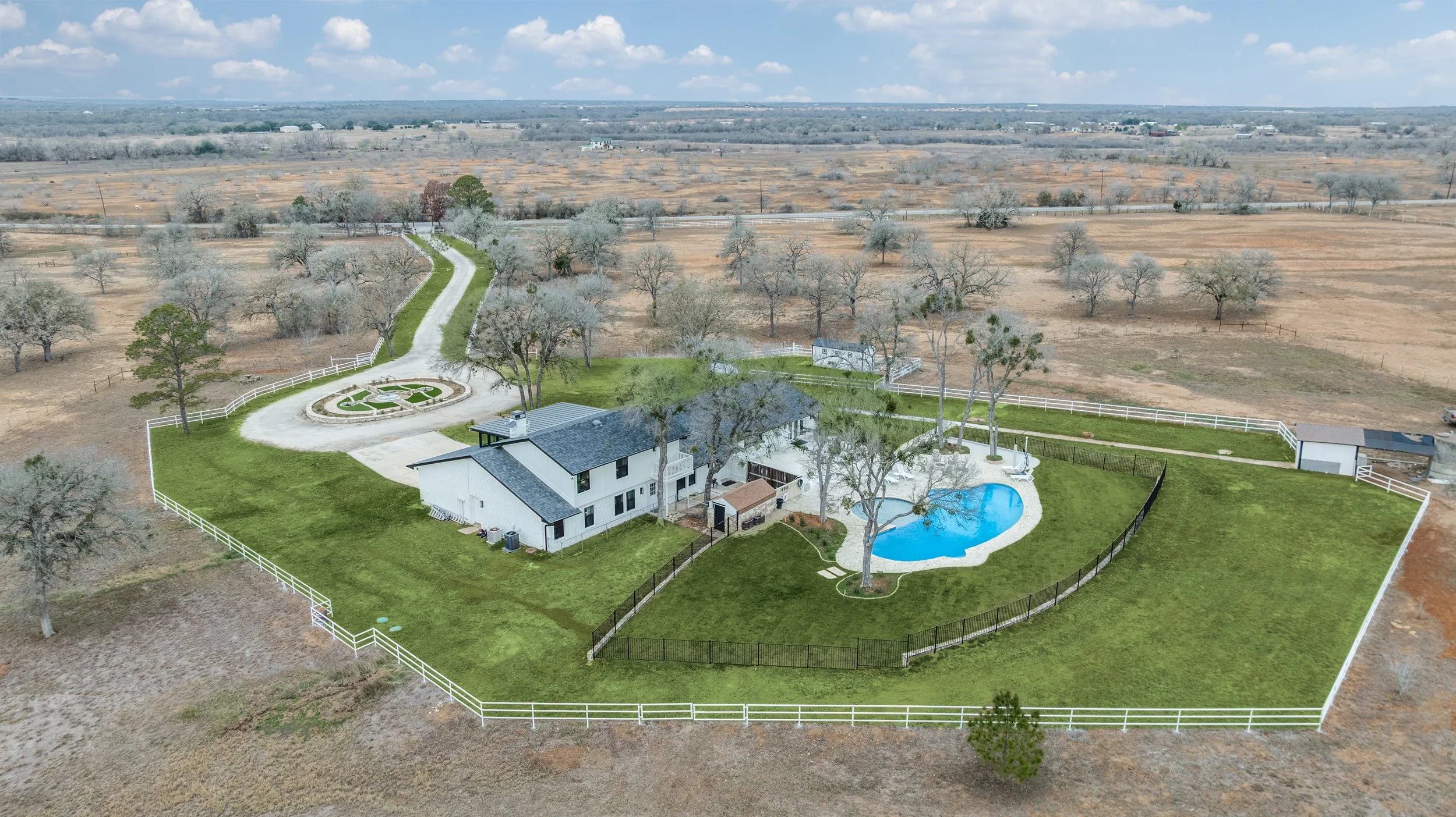 Aerial view of a suburban property with a house, pool, and landscaped yard, surrounded by a white fence, with open fields and farmland beyond.