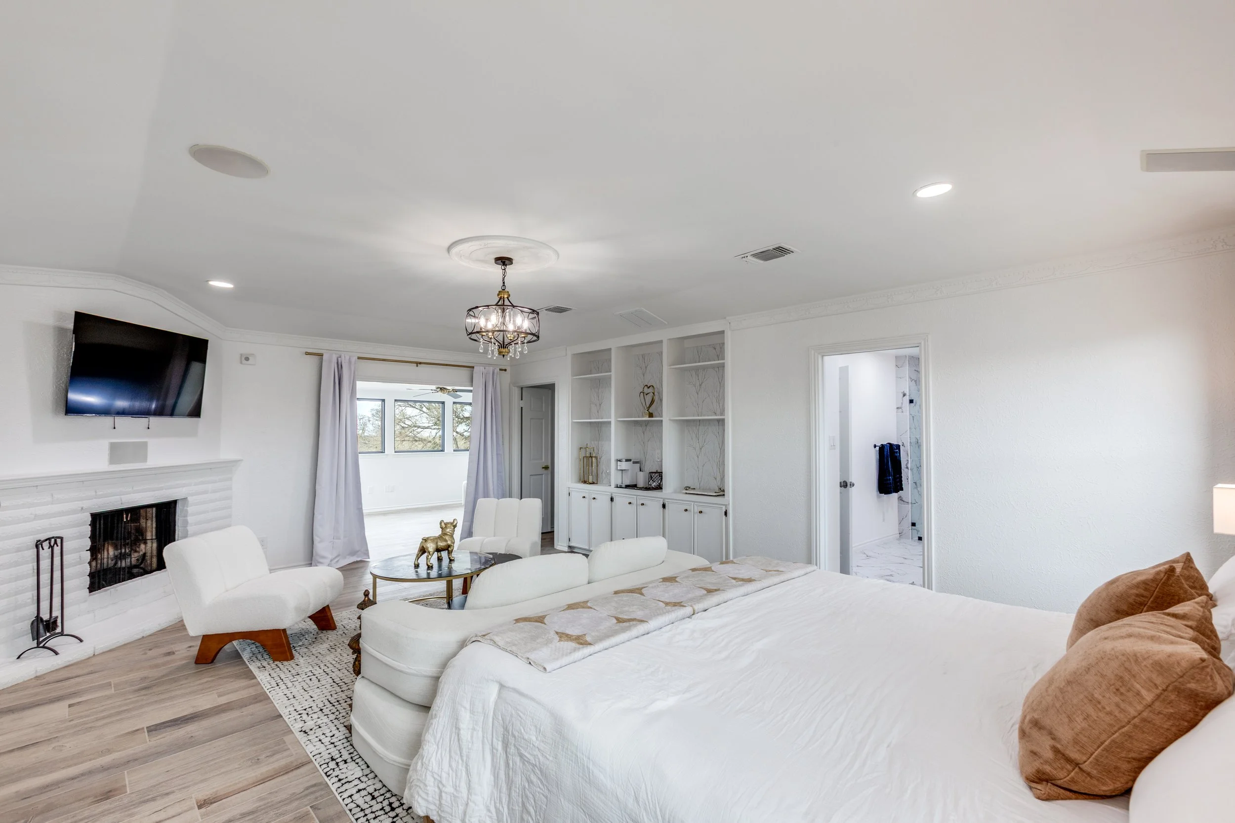 Living room with white walls and ceiling, chandelier, wall-mounted TV, fireplace, windows with white curtains, white furniture including a sofa and chairs, a small round table with a gold dog figurine, and a view into another room with doorway.