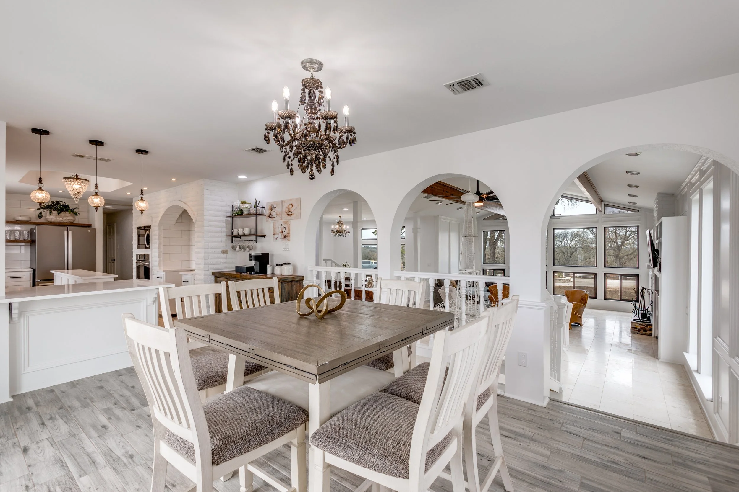 A spacious, bright dining and kitchen area in a modern home. The room features white walls, arched doorways, a large wooden dining table with white chairs, and elegant lighting fixtures including a chandelier and pendant lights. Floor-to-ceiling wind