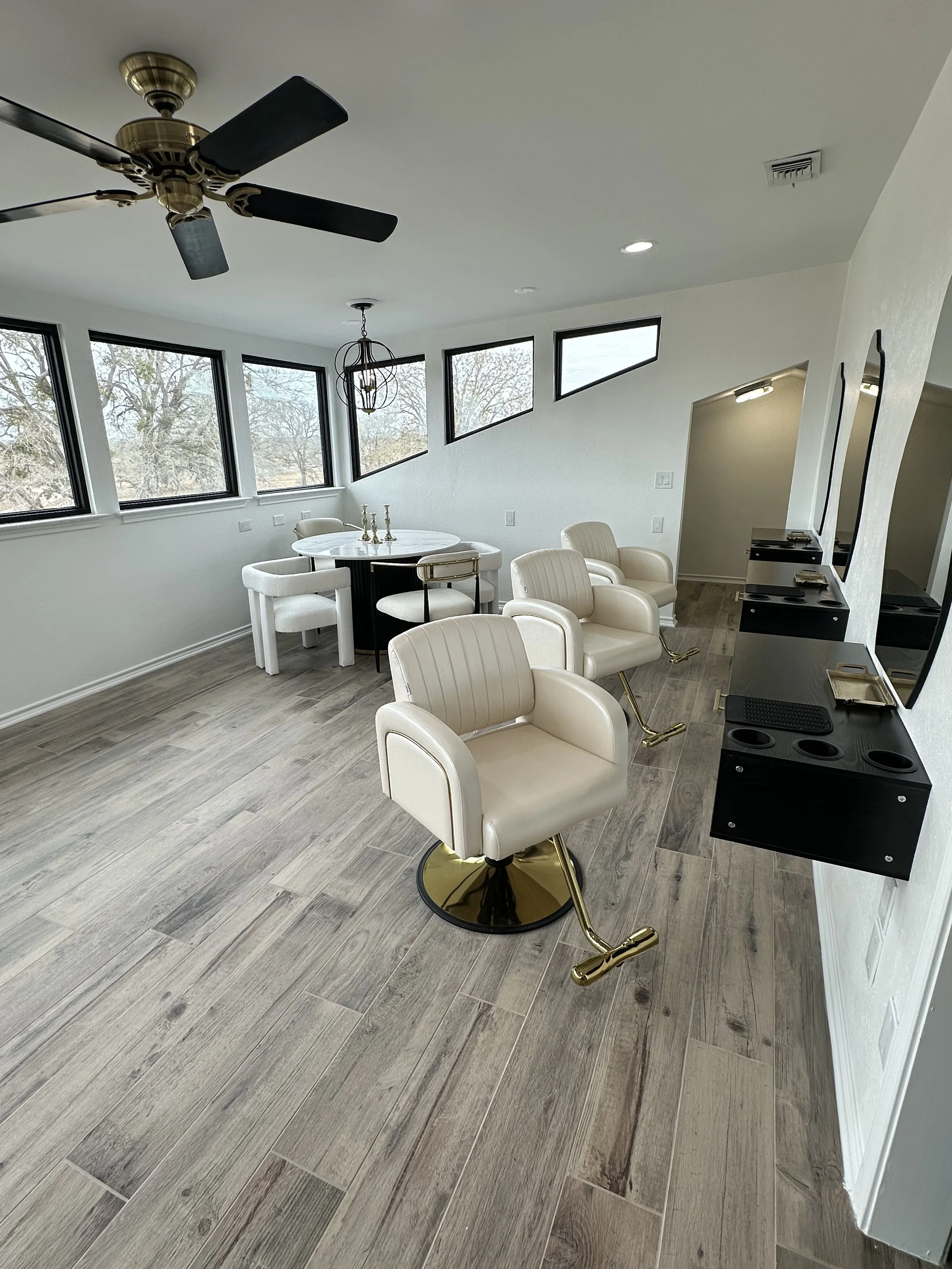 A salon or hair studio with beige barber chairs with gold accents, black and gold styling stations with mirrors, wine-colored flooring, and pastel walls. There are three equal-sized windows, a modern black chandelier, and a ceiling fan.