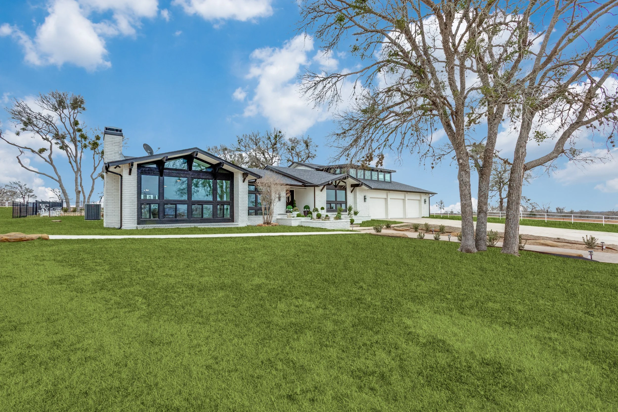 Modern house with large glass windows, white brick walls, surrounded by green lawn and tall trees under a partly cloudy sky.