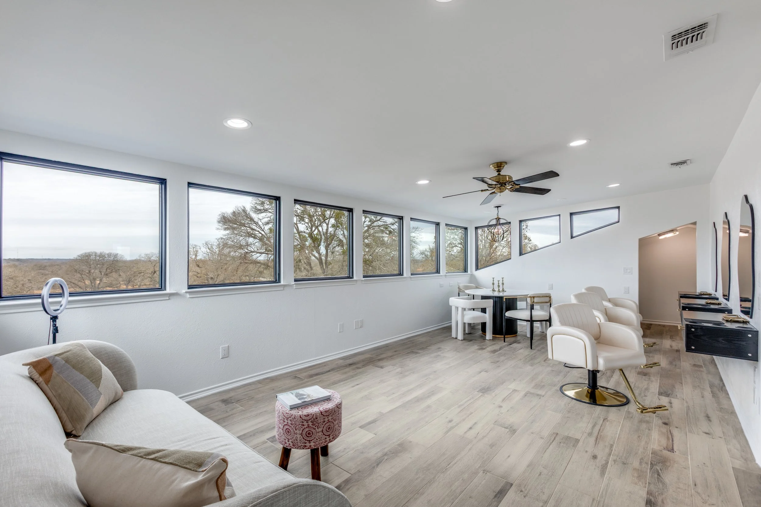 Bright living room with large windows, white walls, a beige sofa with pillows, a pink patterned ottoman, white chairs at a black dining table, modern black and gold chairs, a ceiling fan, and a view of trees outside.
