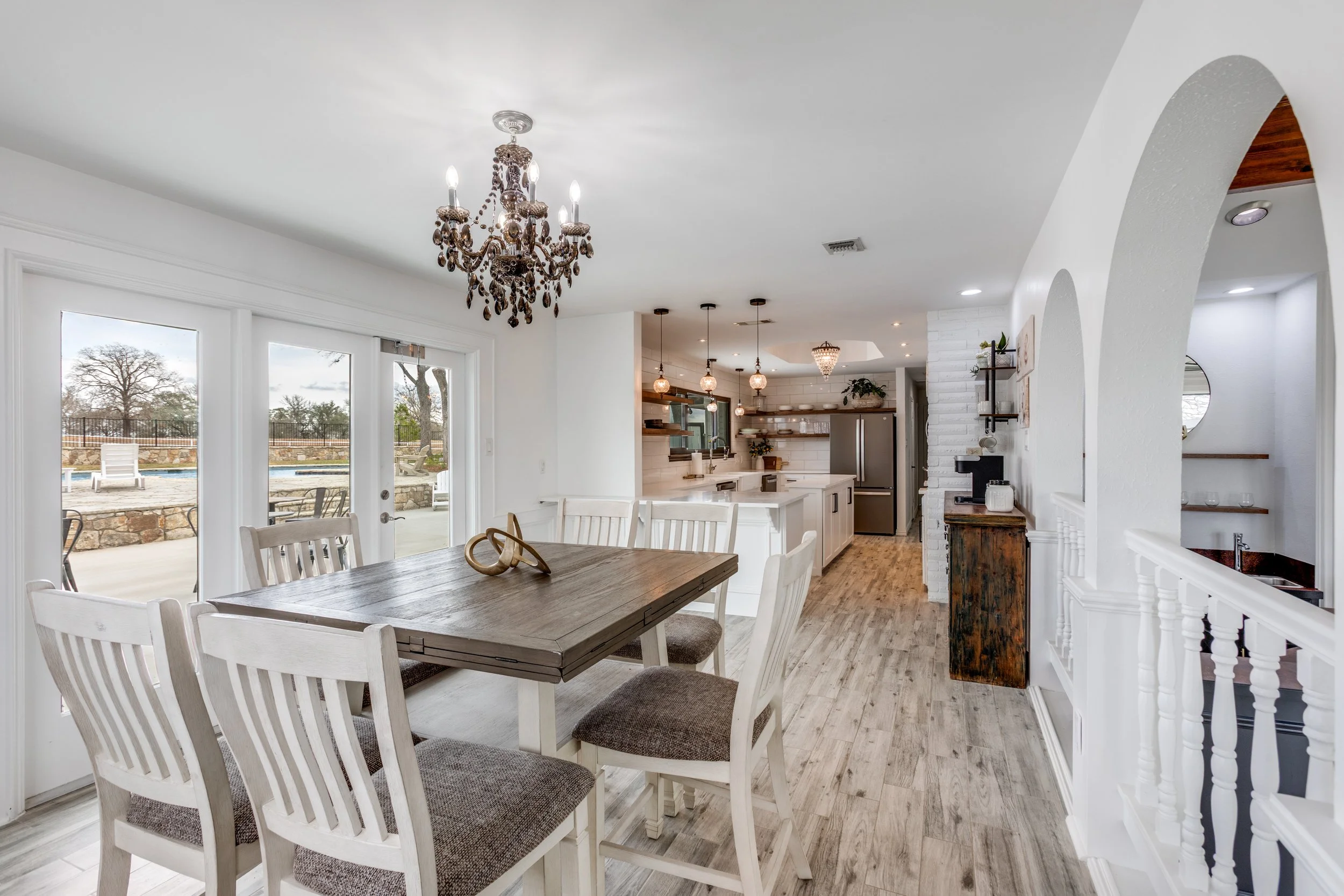 A dining area with a wooden table and white chairs, large glass sliding doors leading outside to a patio and pool area, and a view of a backyard with trees and a fence. The kitchen in the background has hanging pendant lights and white cabinets, and 