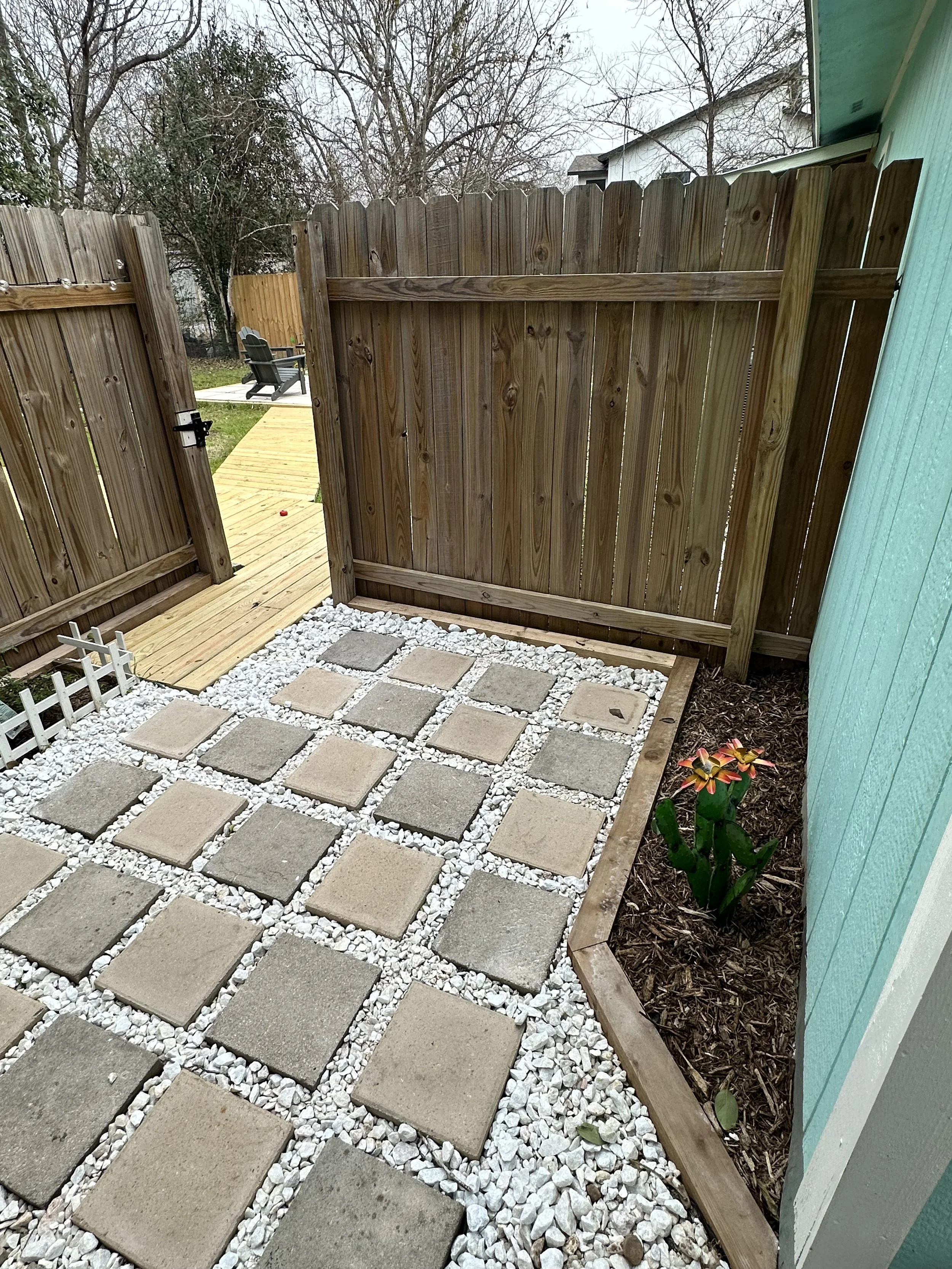 Garden with stepping stones on gravel, a wooden fence, and a flower plant along a turquoise wall.