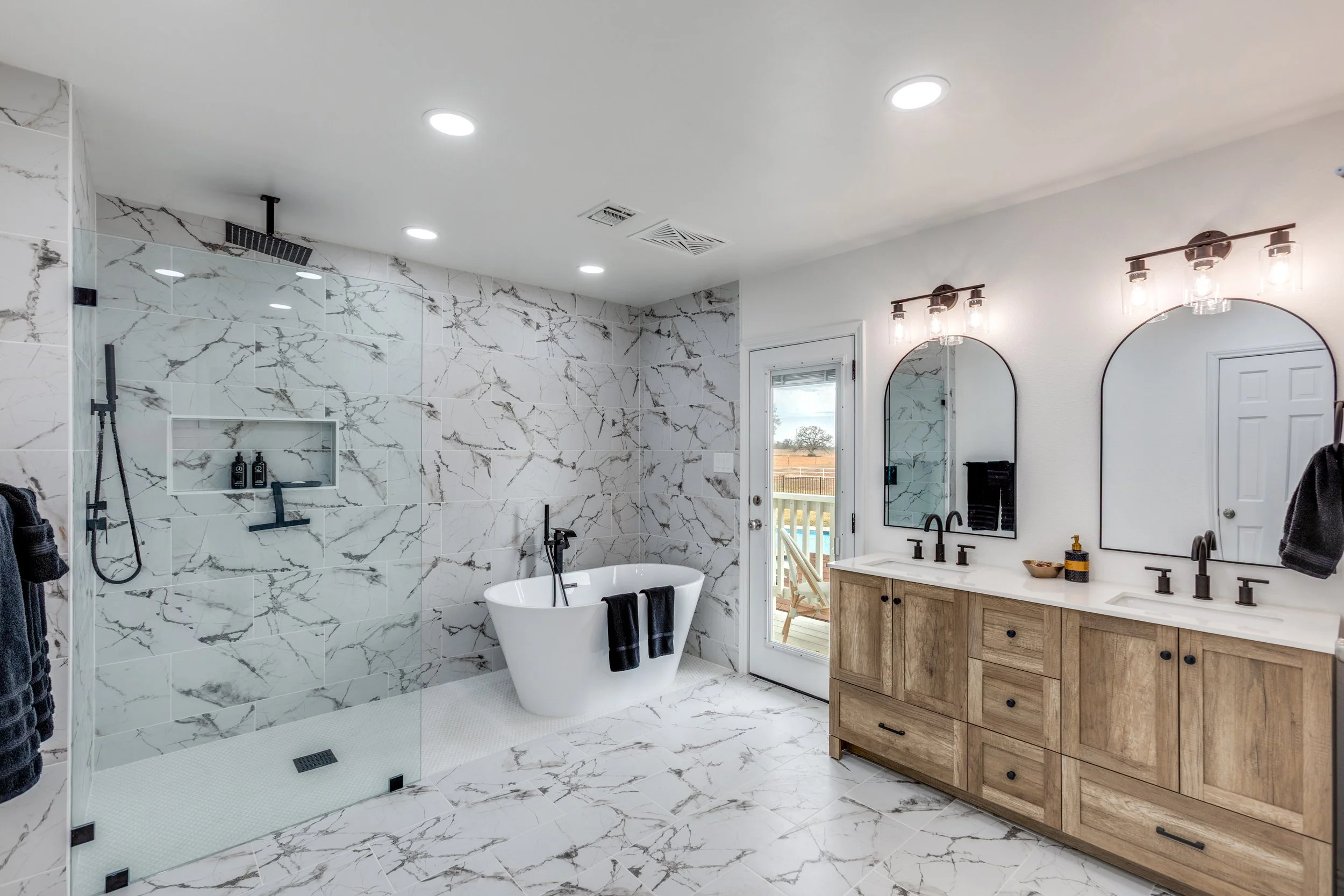 Modern bathroom with white marble walls and floors, a glass-enclosed shower with black fixtures, a freestanding bathtub, a wooden vanity with two mirrors, and a door leading outside to a patio with outdoor furniture.