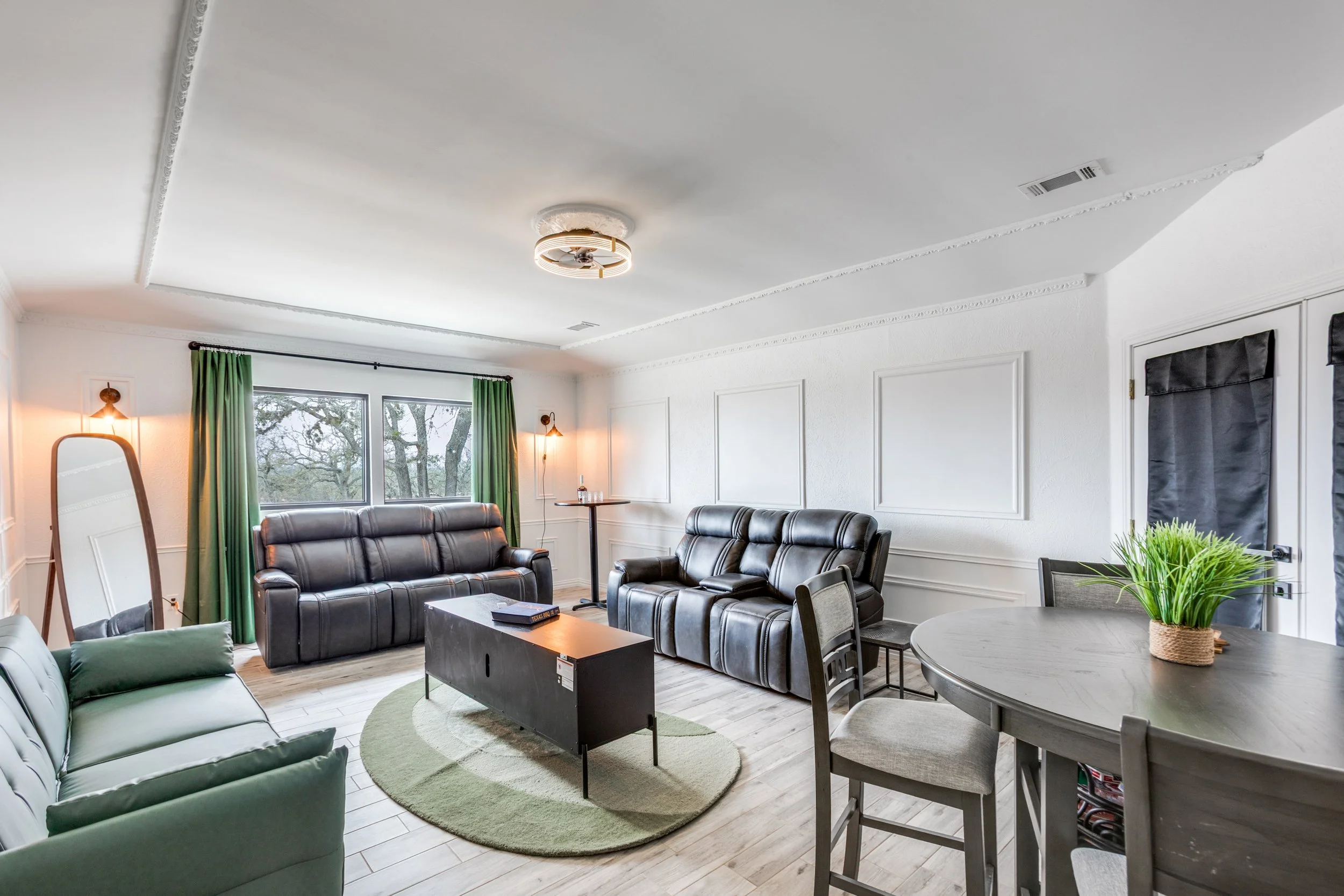 Living room with white walls, large window with green curtains, black leather sofas, green and gray pillows, a round table with a plant, a rectangular table, a mirror, and a ceiling light fixture.