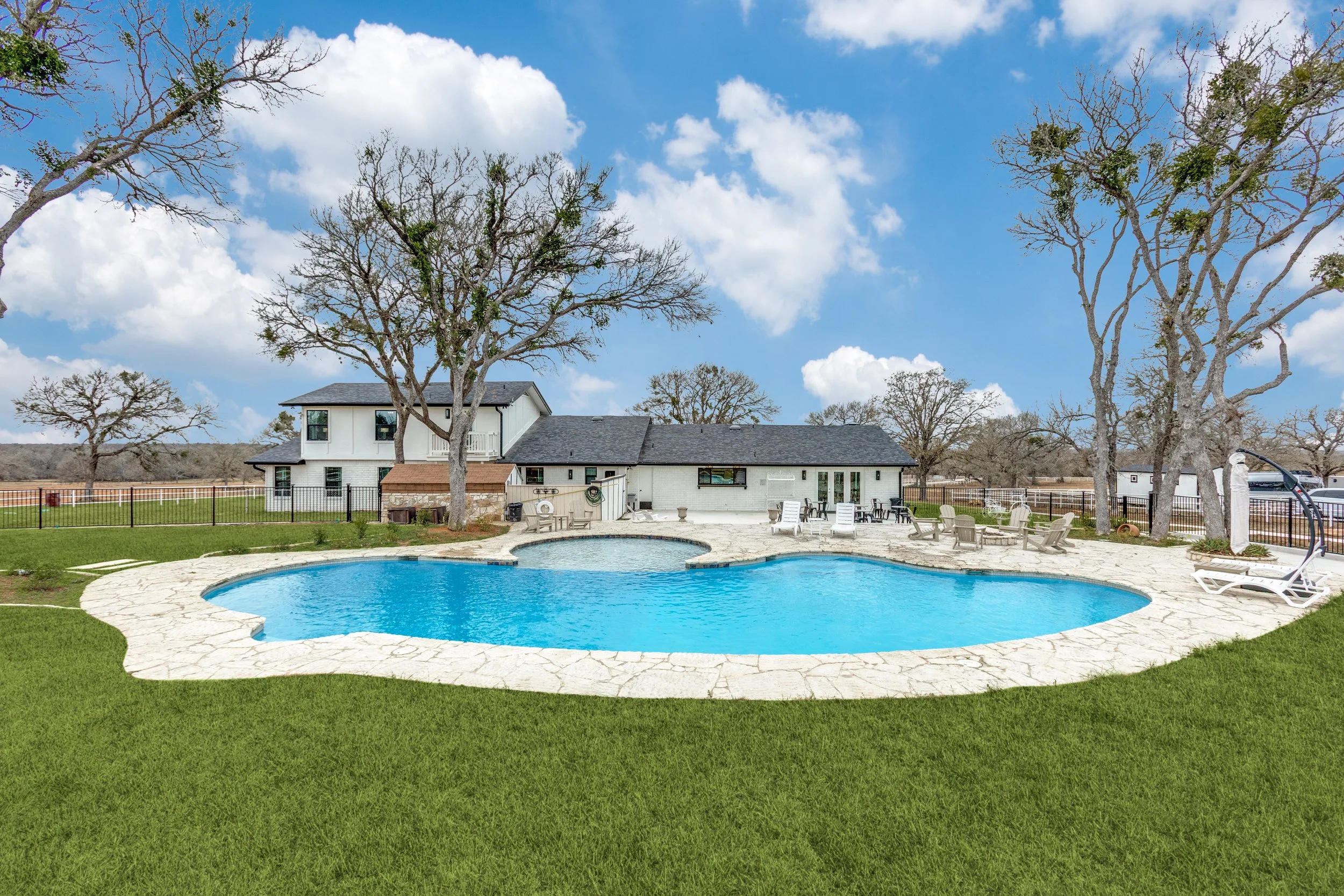 A backyard with a swimming pool surrounded by a stone patio, outdoor furniture, and a white house with a gray roof. There are tall leafless trees and a blue sky with clouds.