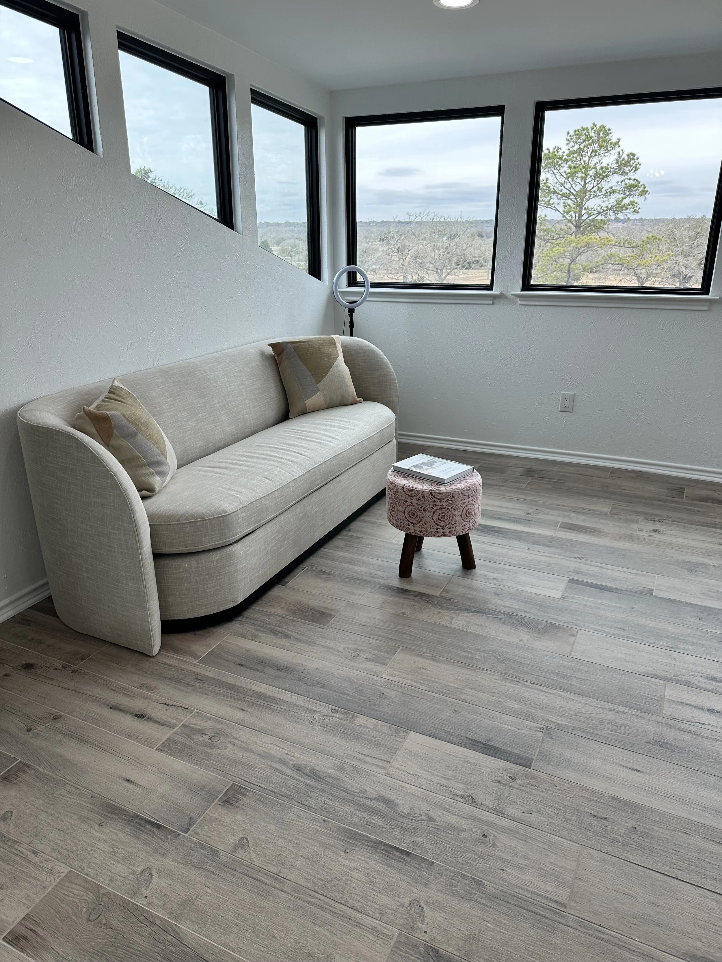 A beige sofa with two matching pillows, a small pink patterned ottoman, and a ring light beside the sofa, in a room with large windows showing an outdoor view of trees and cloudy skies.