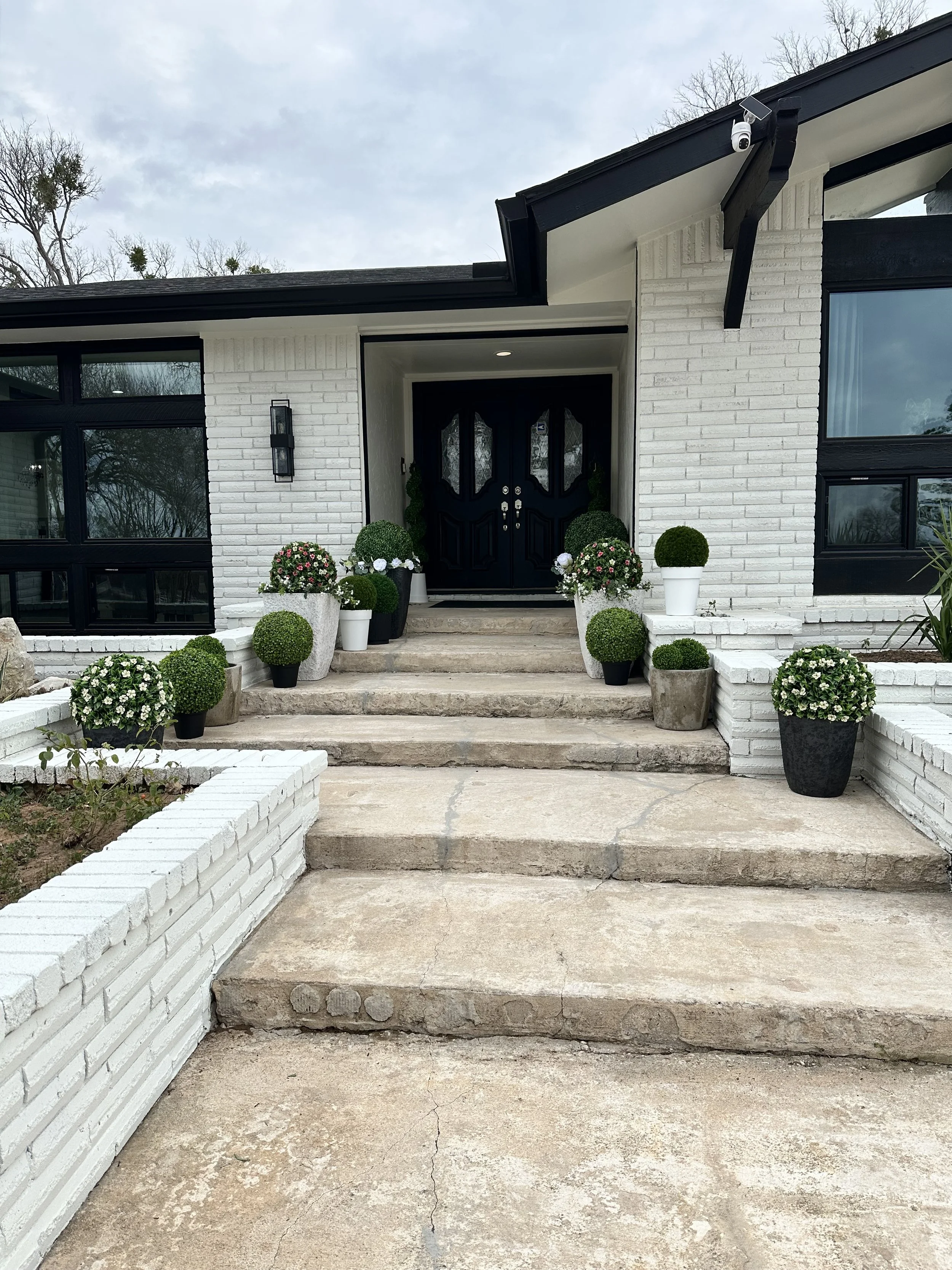 Front steps leading to a black front door decorated with potted plants and flowers, white brick exterior, and large windows.