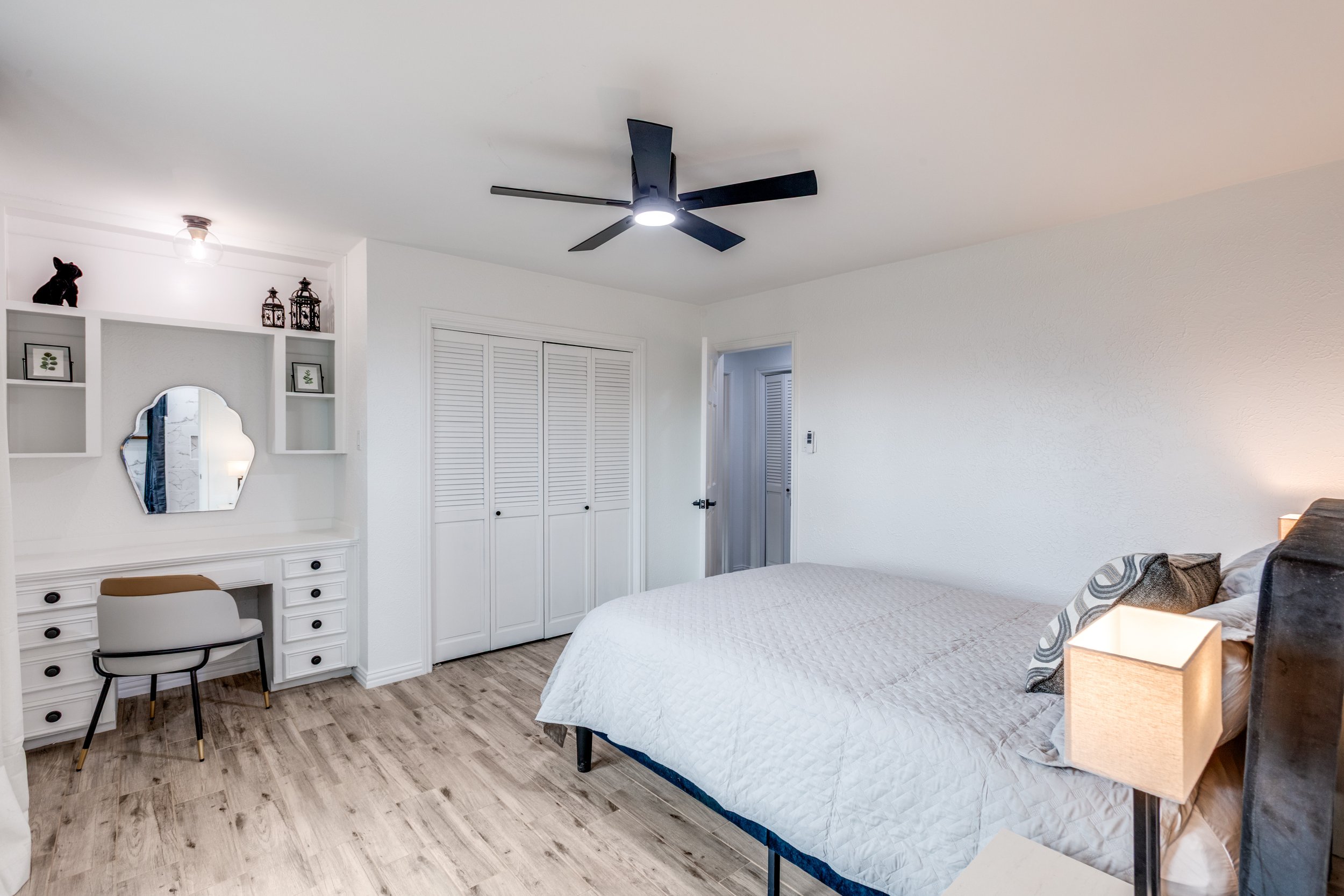 A bedroom with a white bed, pillows, and bedside table on a wood floor. A ceiling fan with lights is on the ceiling. There is a white closet with louvered doors, a small desk with a chair, and decorative items on shelves.