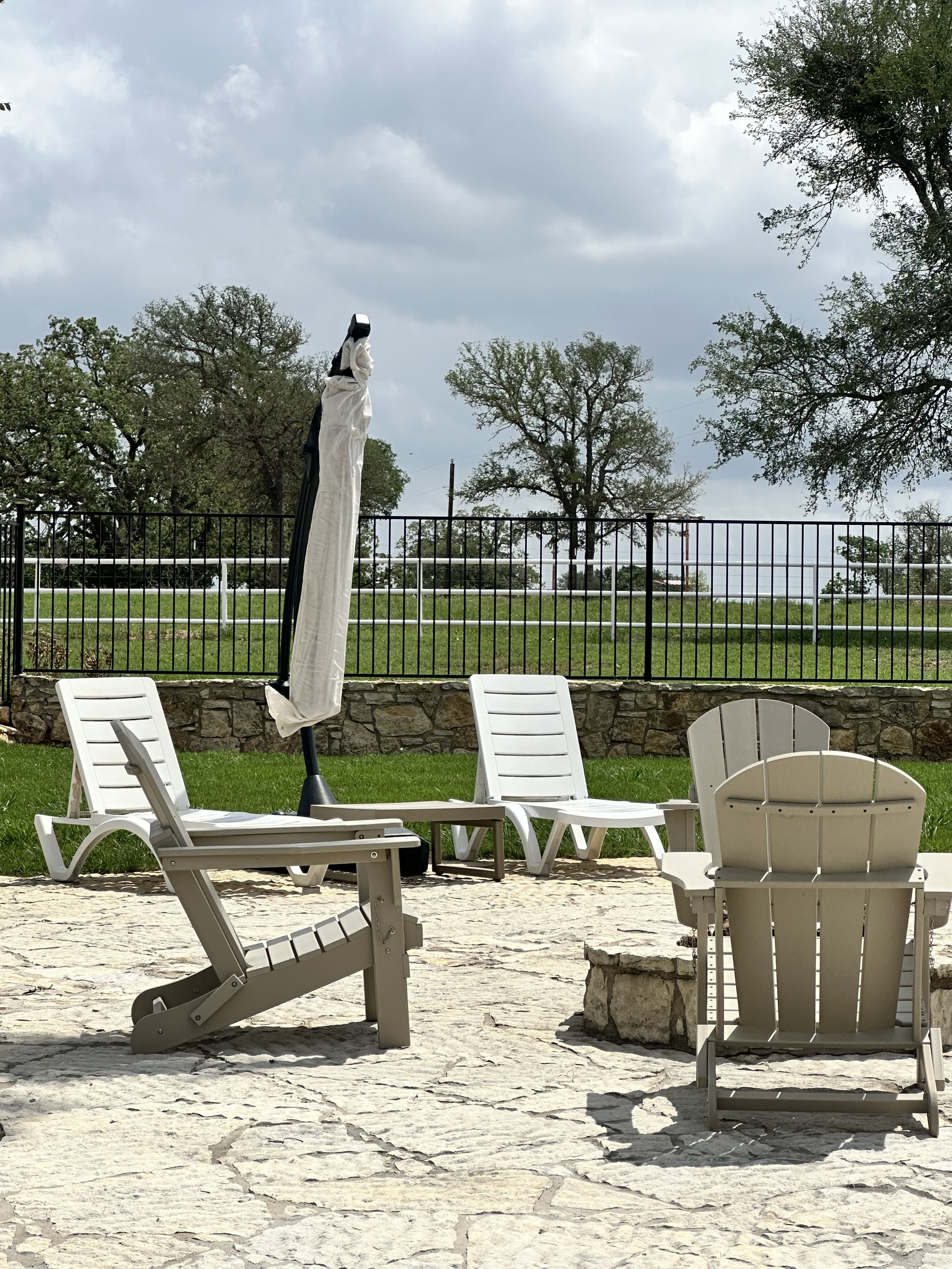 Empty plastic chairs and a closed patio umbrella on a stone patio in a backyard, with a green lawn, a black metal fence, and trees in the background under a cloudy sky.