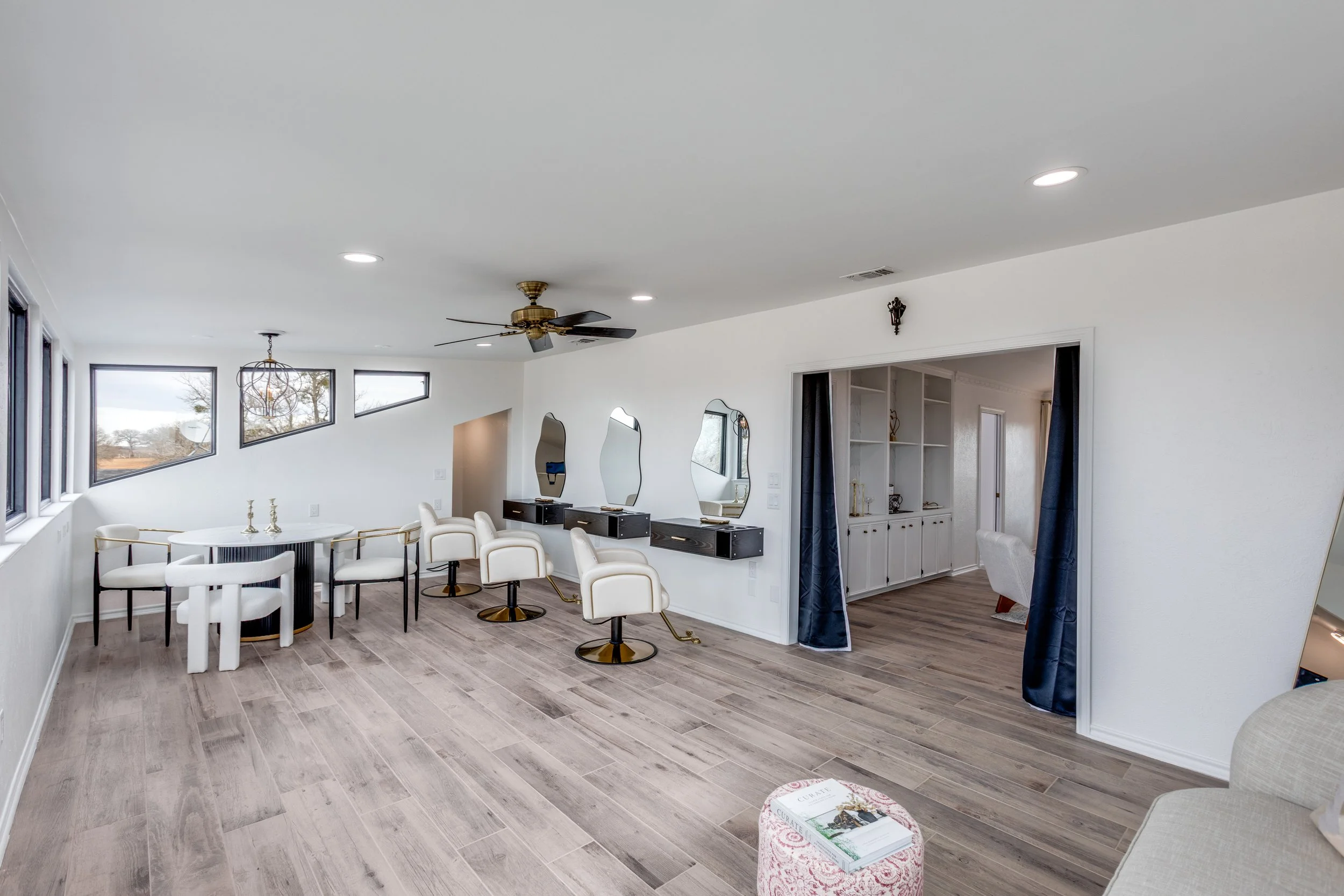 A living room and dining area with white walls, gray wood floors, and large windows letting in natural light. There are white chairs at a round dining table, a black and white salon station with mirrors, and decorative curtains. Ceiling lights and a 