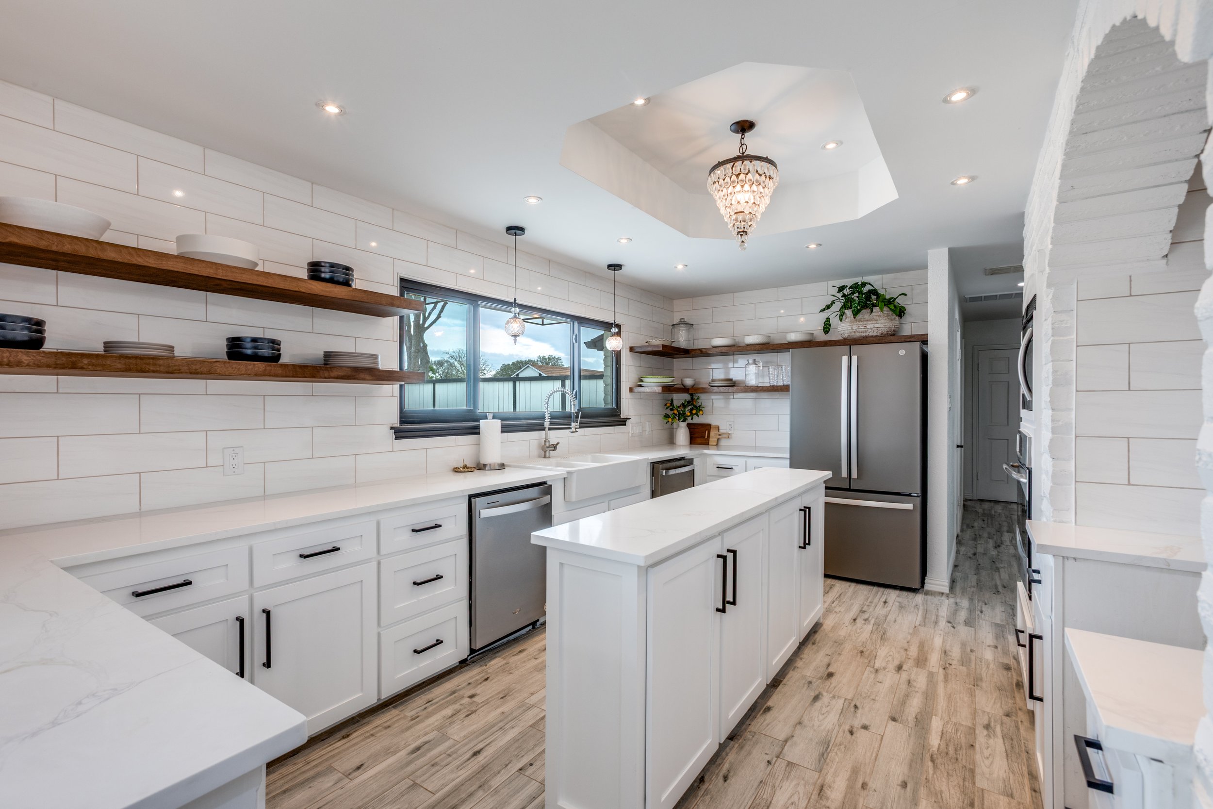 Modern kitchen with white cabinets, open wooden shelves with dishes, large window, and a chandelier hanging from the ceiling.