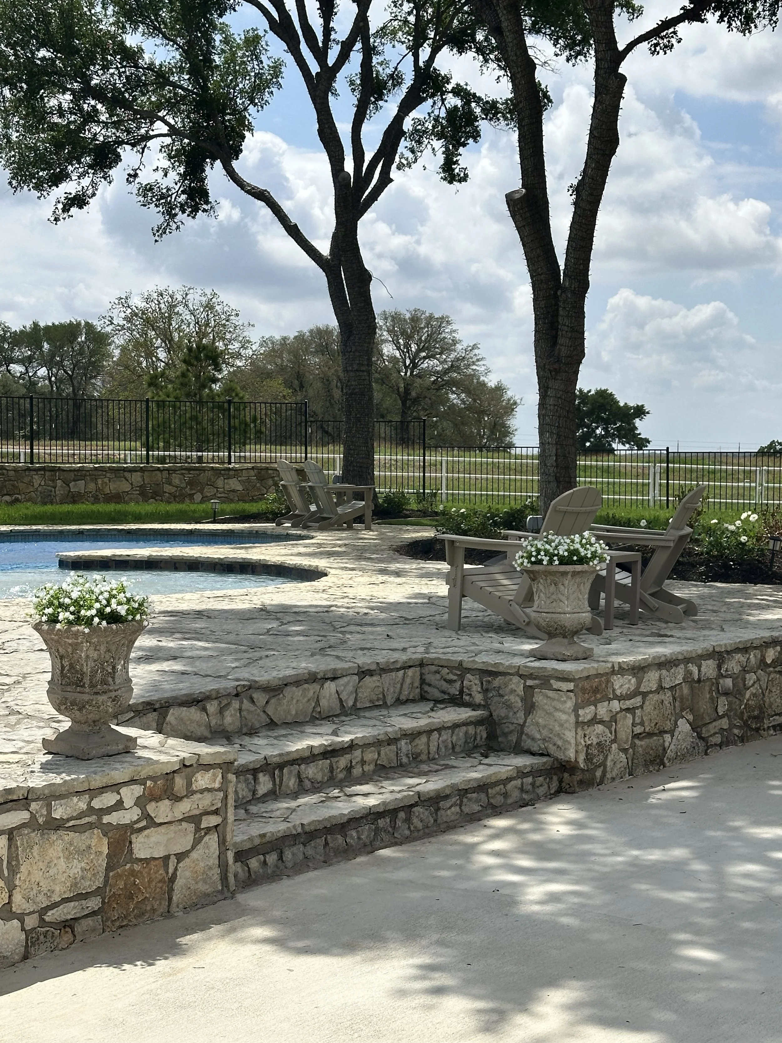 A backyard scene with a stone patio, wooden chairs, white potted flowers, tall trees, a pool, and a grassy fence line under a partly cloudy sky.