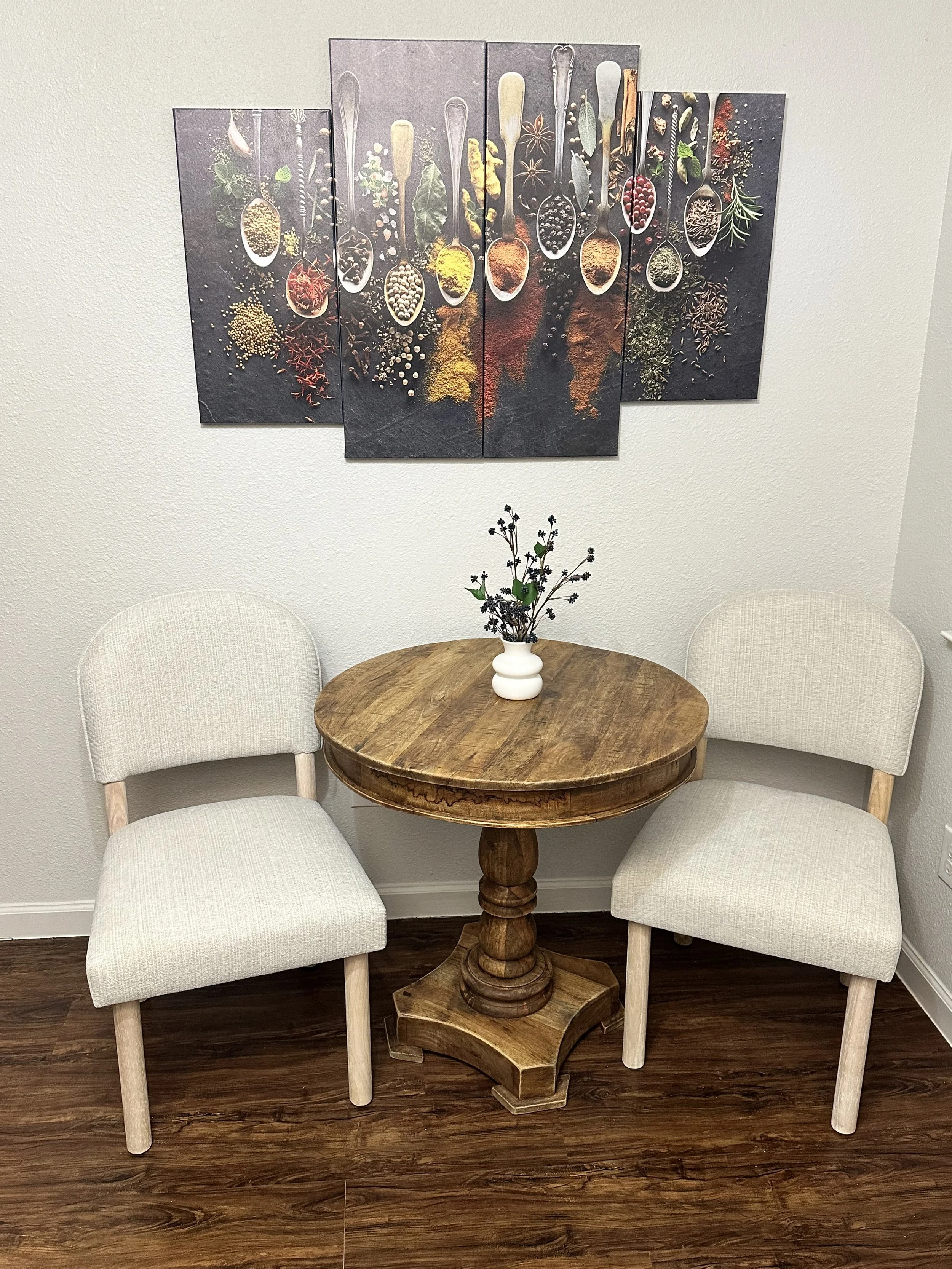 A small round wooden table with a white ceramic vase of black and green dried flowers, surrounded by three off-white upholstered chairs with light wooden legs, and a multi-panel wall art piece with various spices and herbs displayed in spoons, hangin