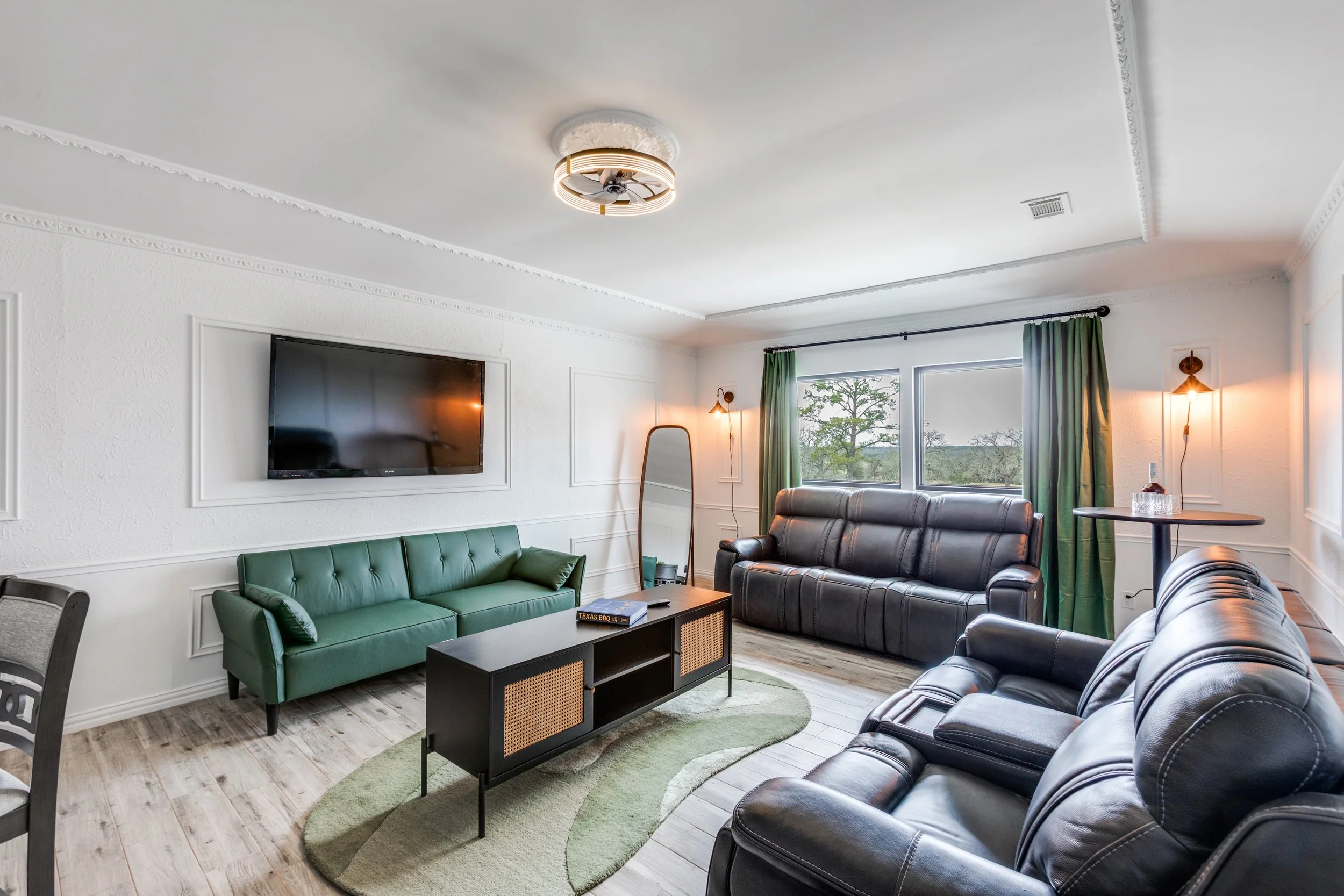 A living room with a black leather sofa, a green sofa, a wall-mounted TV, a coffee table with a book, large window with green curtains, wooden flooring, decorative wall lights, and a modern ceiling fan.
