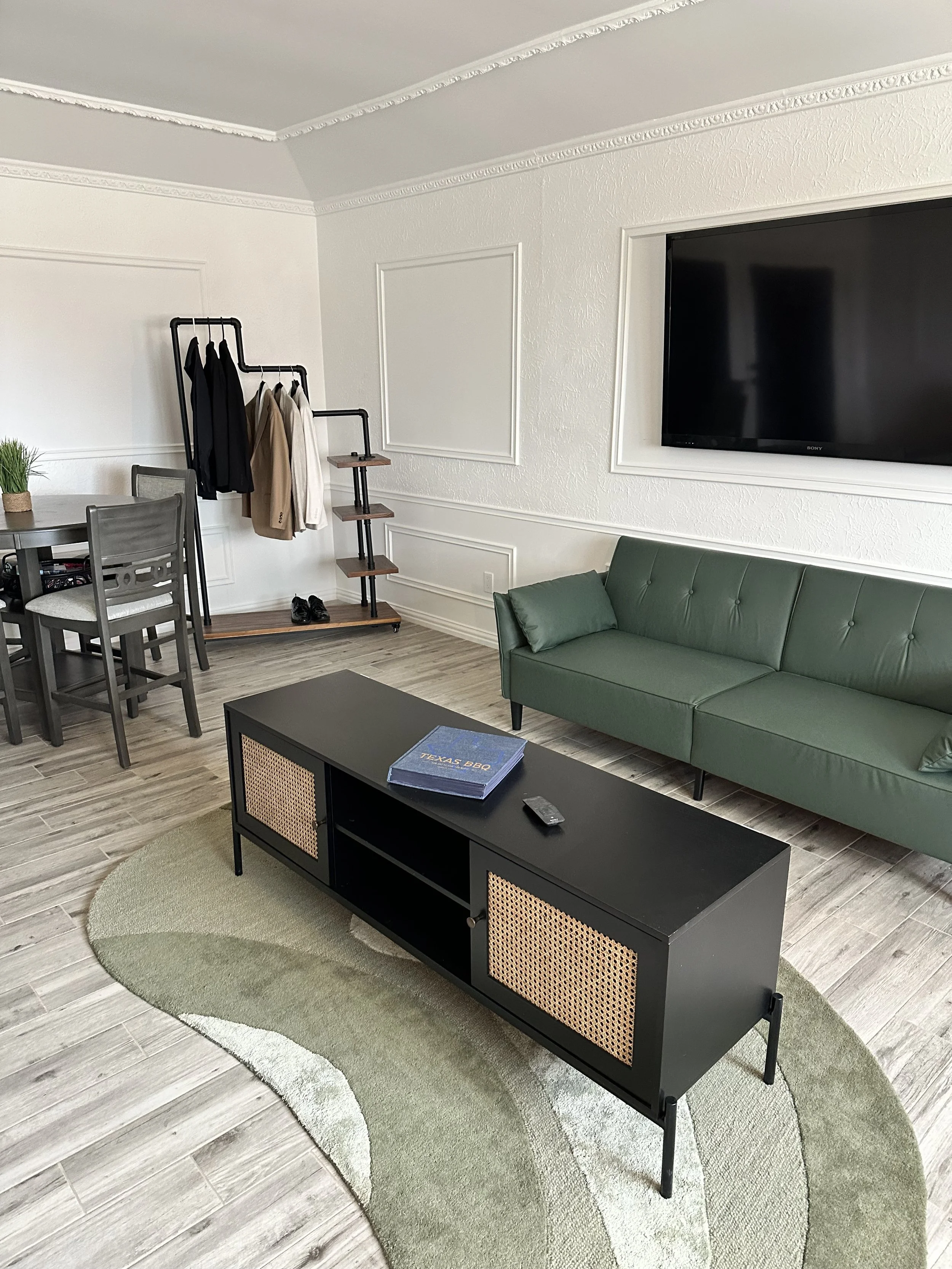 A living room with a green couch, a black coffee table, and a black TV mounted on a white wall. There is a black clothing rack with jackets and shirts, a small wooden shelving unit below it, and a dining table with chairs. A round green rug on light 