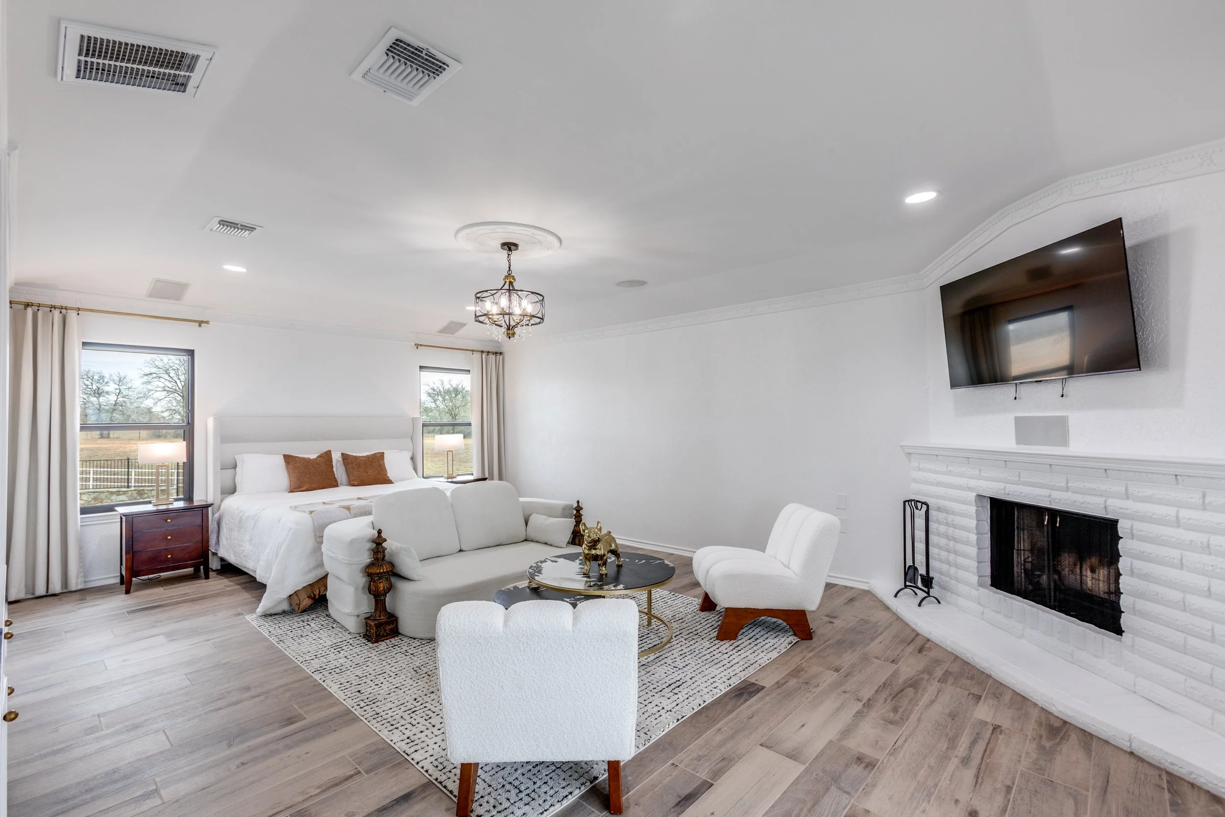 A bright, modern living room with a white fireplace, flat-screen TV, and large windows with white curtains. The room features a white sofa, a white armchair, a black and gold coffee table, and a chandelier with multiple lights. The walls are white, a