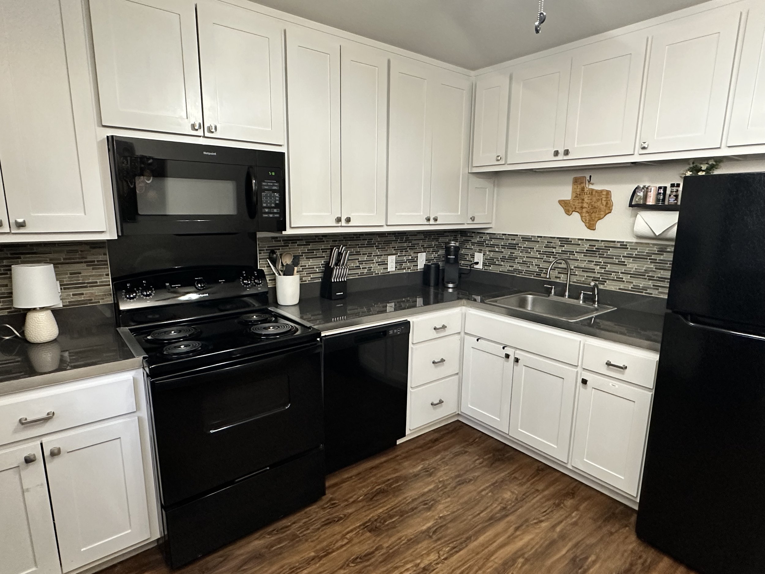 Kitchen with white cabinets, stainless steel sink, black stove, black microwave, and wood flooring, with items on countertops and a wall decoration.