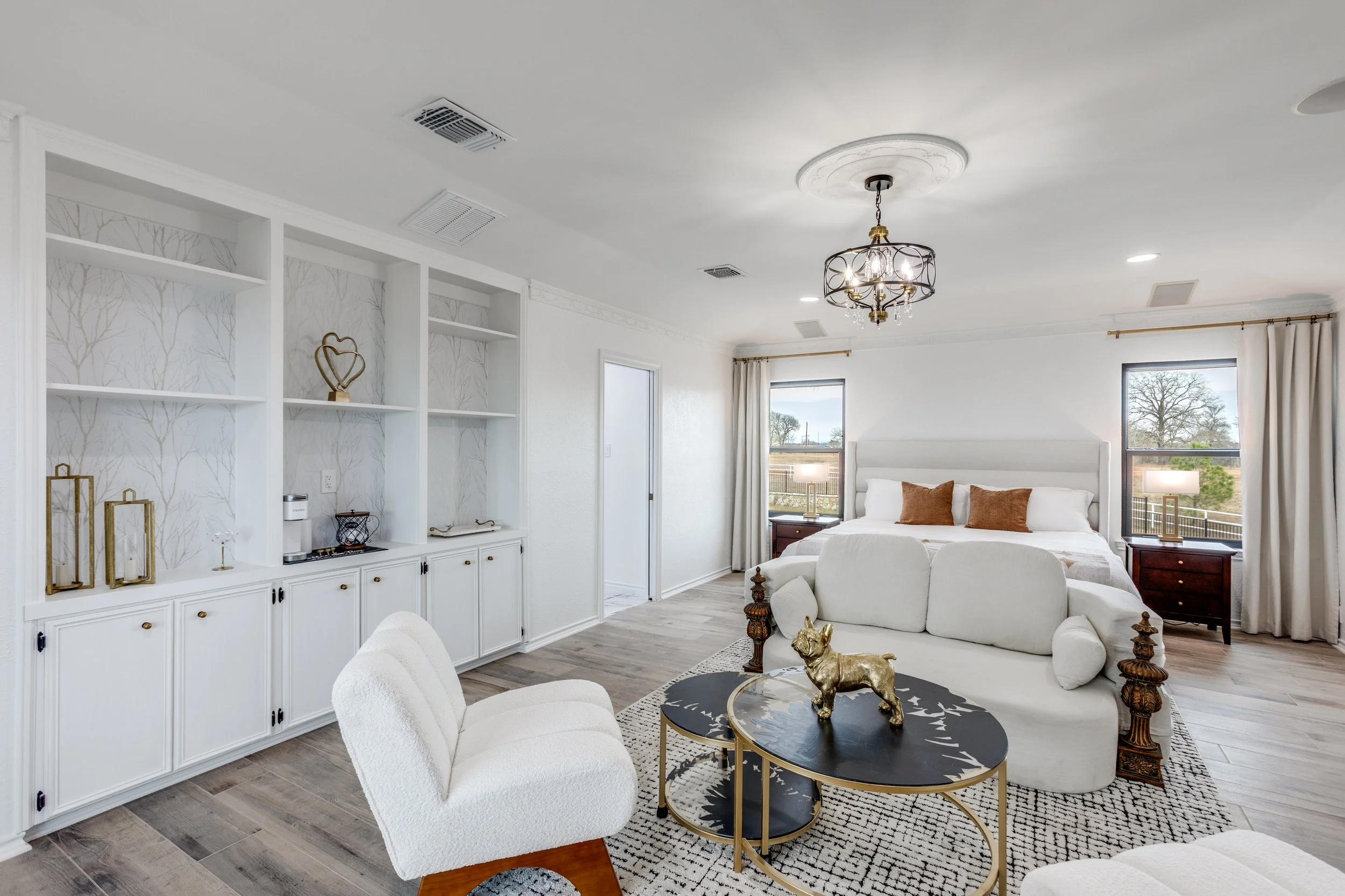 Bright modern bedroom with white walls, large windows, a chandelier, a white bed with brown pillows, a white armchair, dark wooden nightstands, and decorative gold accents on a black and gold coffee table with a dog sculpture.