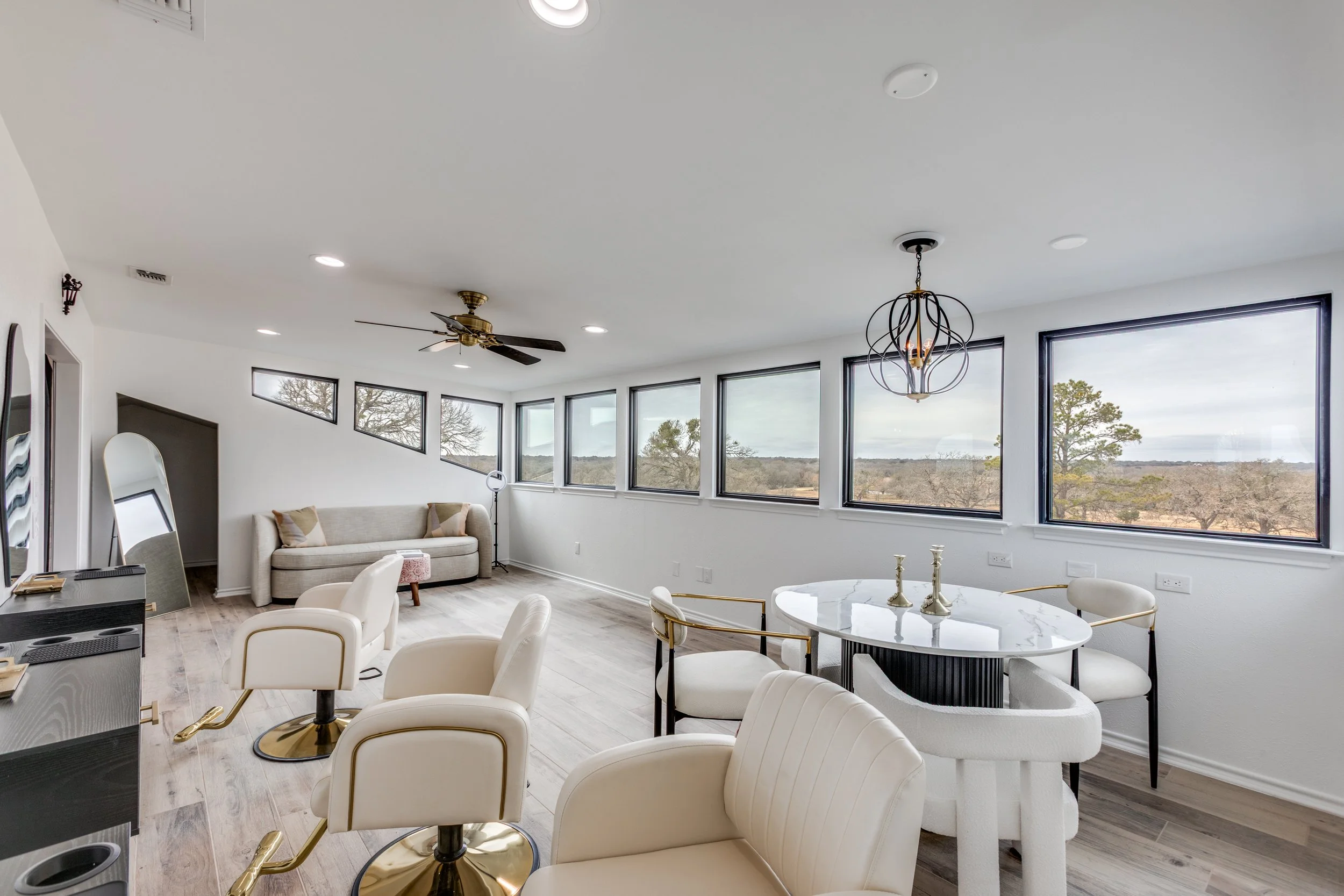Bright living room with large windows showing an outdoor landscape, white walls, modern chandelier, ceiling fan, white and gold chairs around a round marble table, beige sofa with throw pillows, and a coffee table.