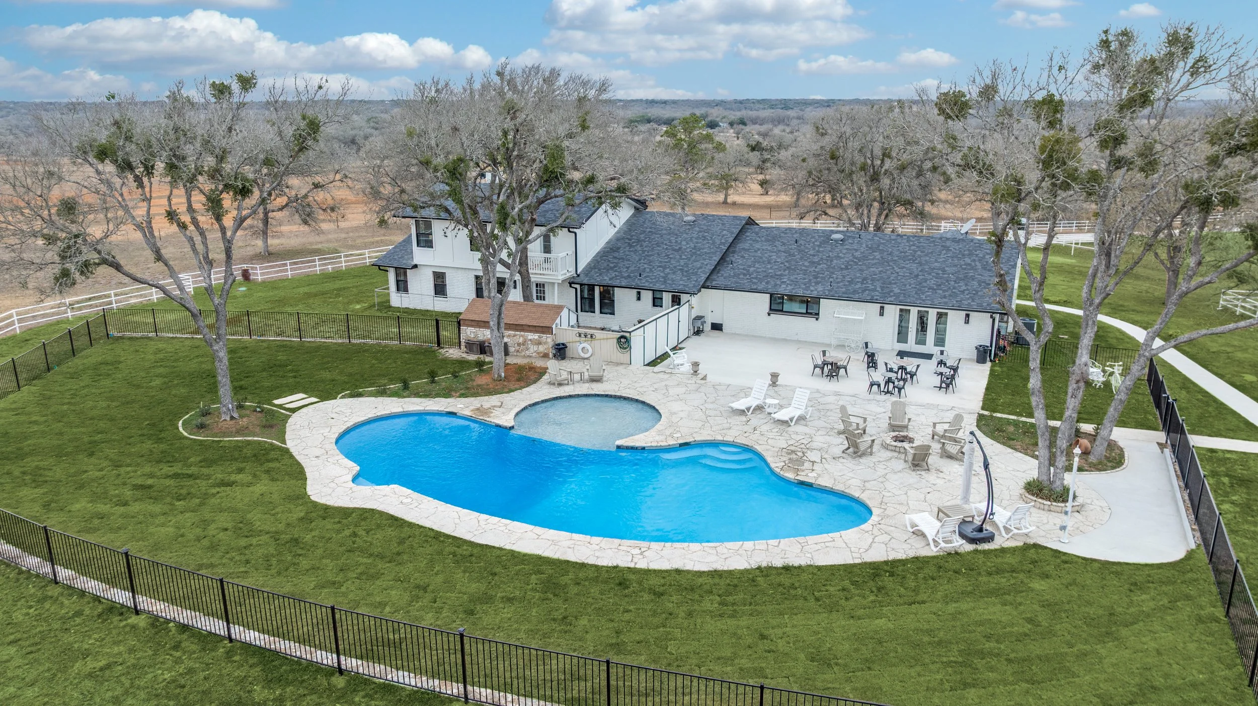 A backyard with a kidney-shaped swimming pool, surrounding concrete patio with outdoor furniture, barbecue area, and a white house with a gray roof. Tall trees and a white fence enclose the yard, with open land and cloudy sky in the background.