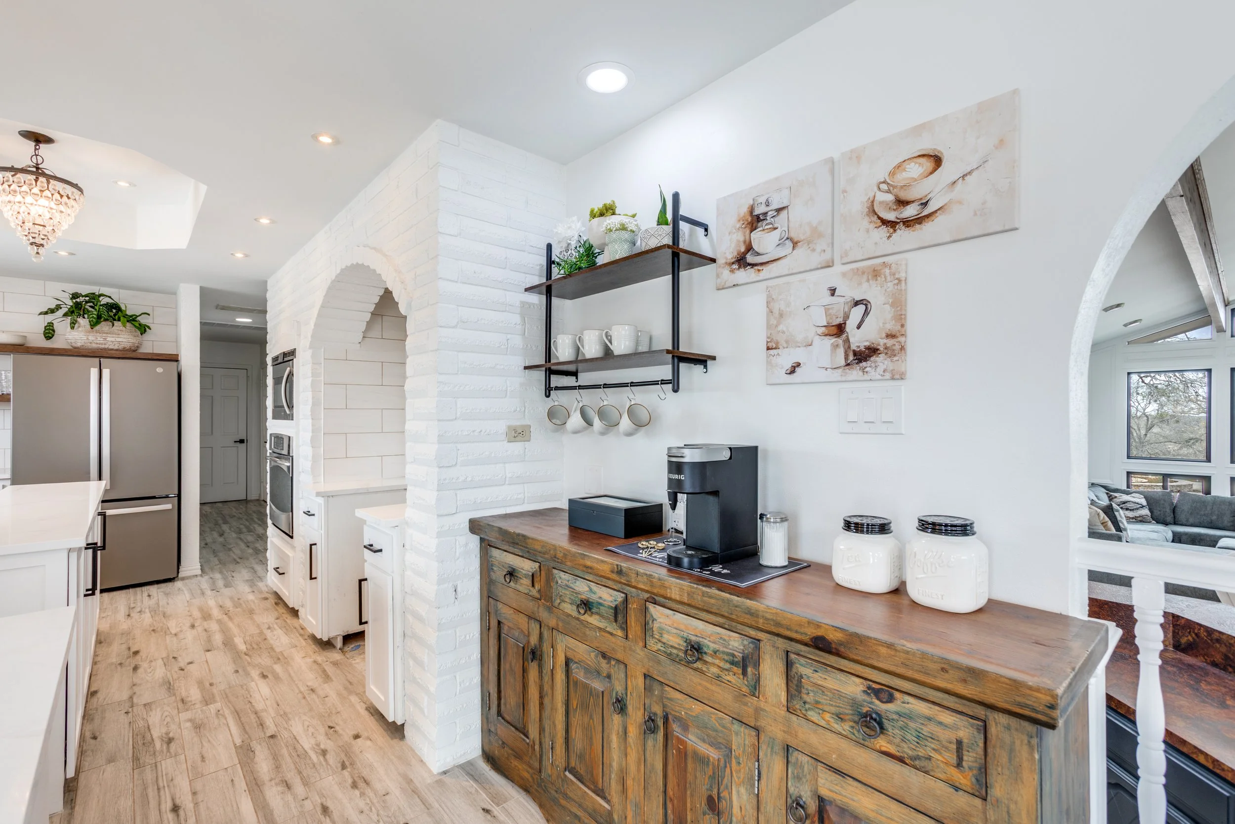 Interior view of a kitchen with white brick and walls, a wooden sideboard with coffee mugs, jars, and a coffee machine, and wall art featuring coffee cups and beans.