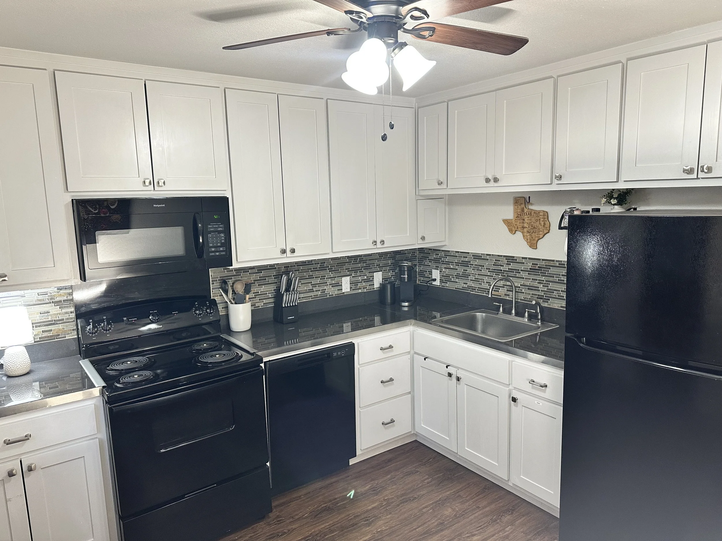 A modern kitchen with white cabinets, black appliances, and a black countertop. There is a mounted microwave oven above a stove, a sink under a window, small appliances on the counter, and decorative items on the wall.