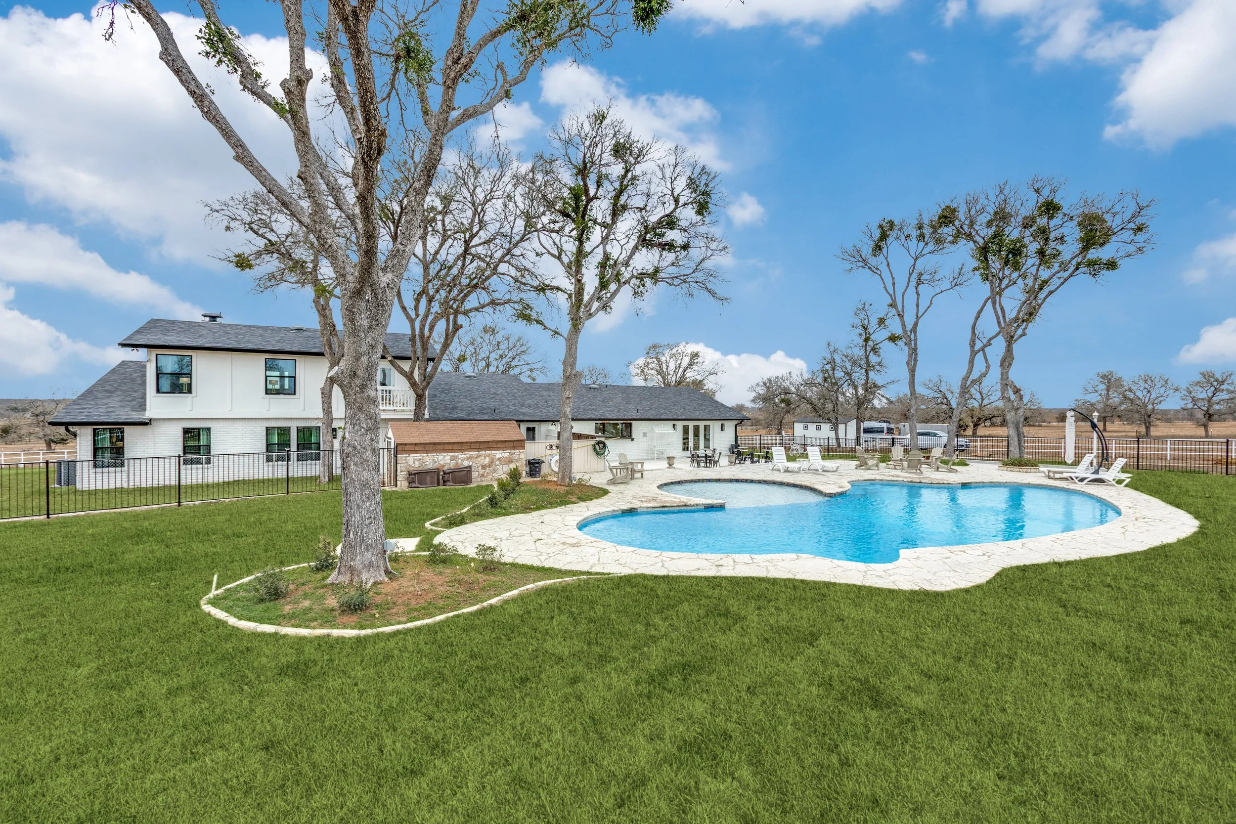 A backyard with a swimming pool, several lounge chairs, a house with a black roof, leafless trees, and a blue sky with clouds.