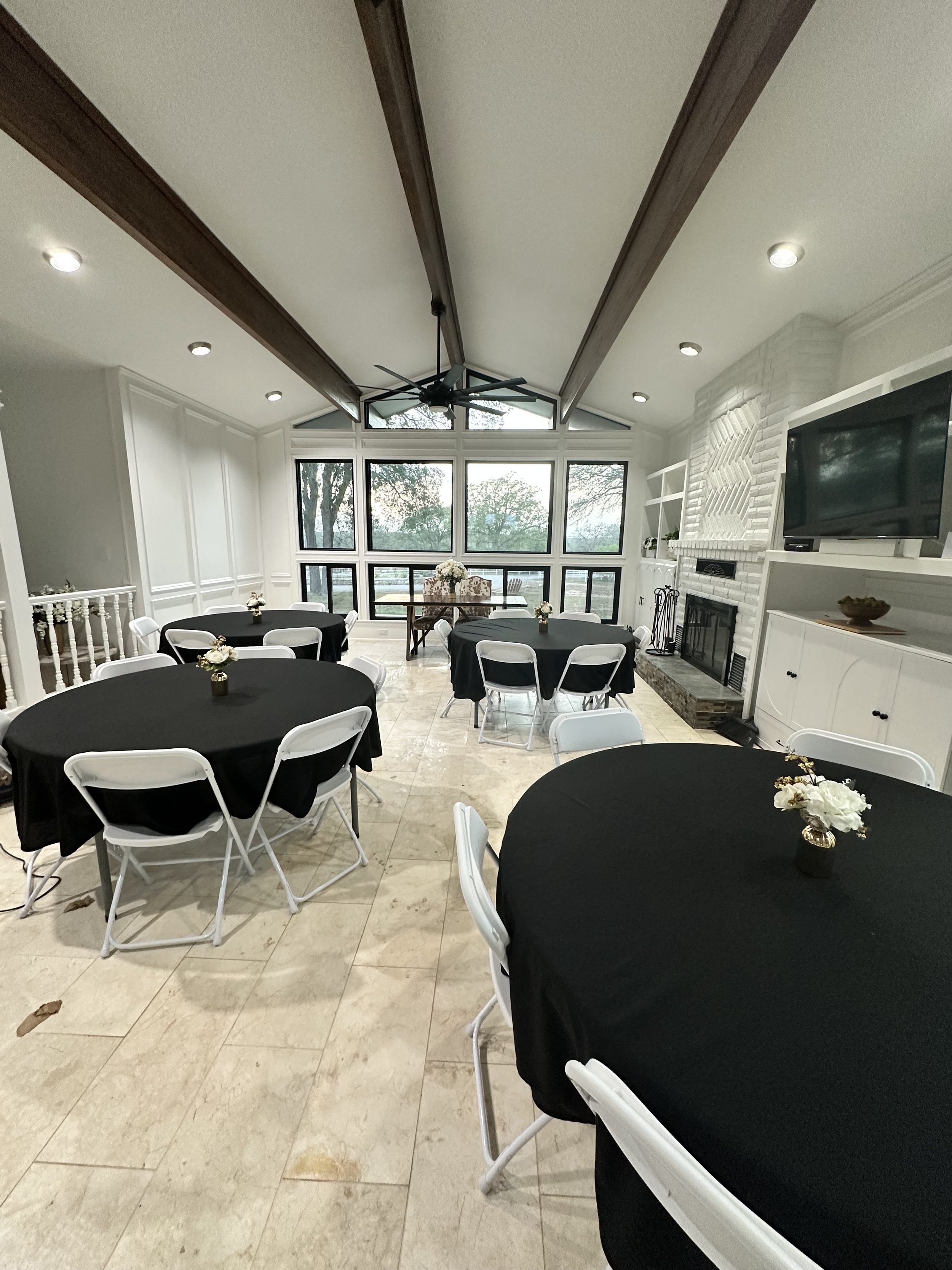 A decorated indoor space with black covered round tables and white folding chairs, each table having a small floral centerpiece, large windows with trees outside, a white brick fireplace, and a ceiling fan.