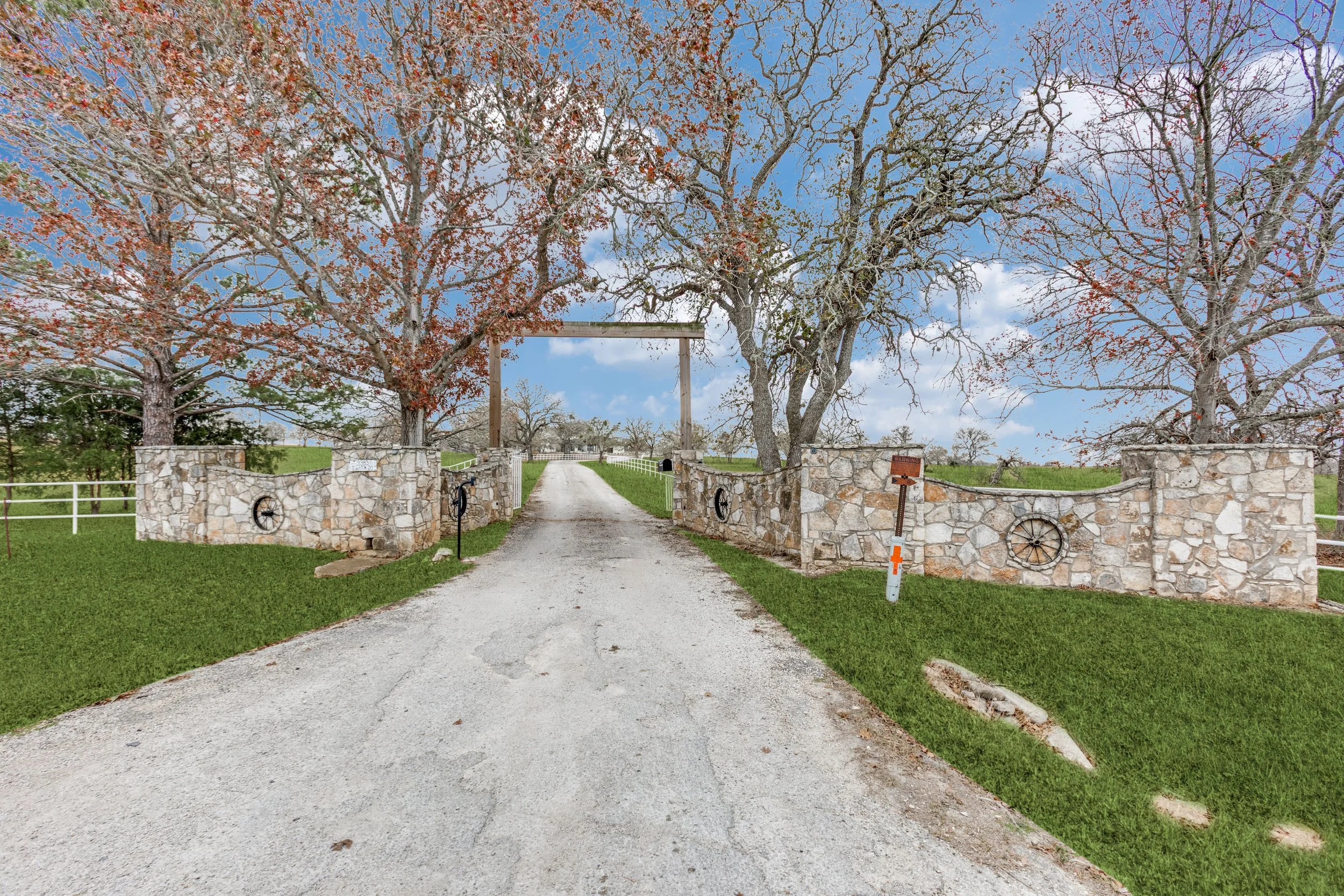 A rural driveway lined with stone wall fencing and large leafless trees on either side, leading to a wooden gate with a blue sky and scattered clouds above.