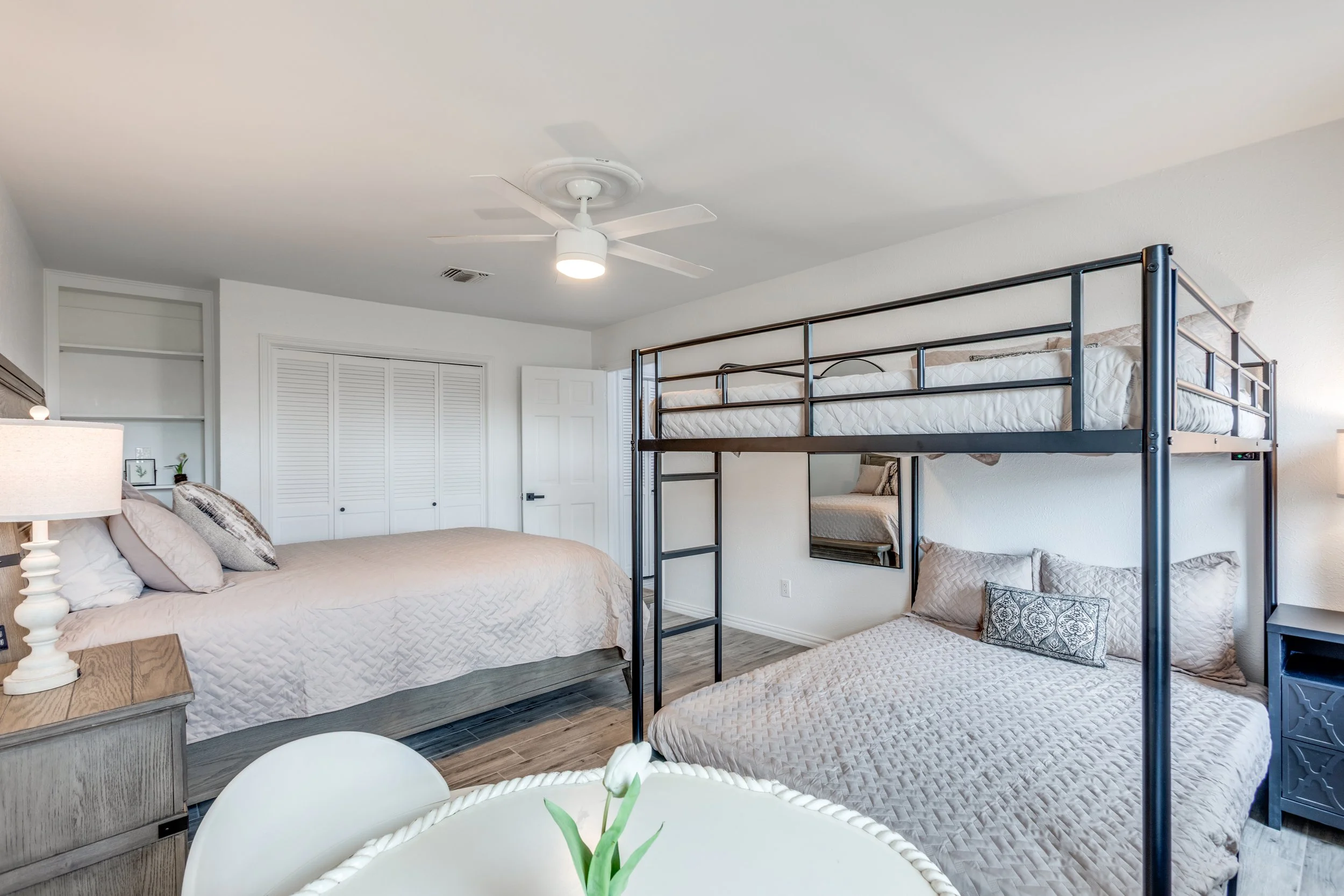 A bedroom with two beds, one king-sized with beige bedding and pillows, and a metal bunk bed with a twin mattress, a decorative pillow, and a window behind it. The room has white walls, a white ceiling fan with a light, a dresser with a lamp, a small