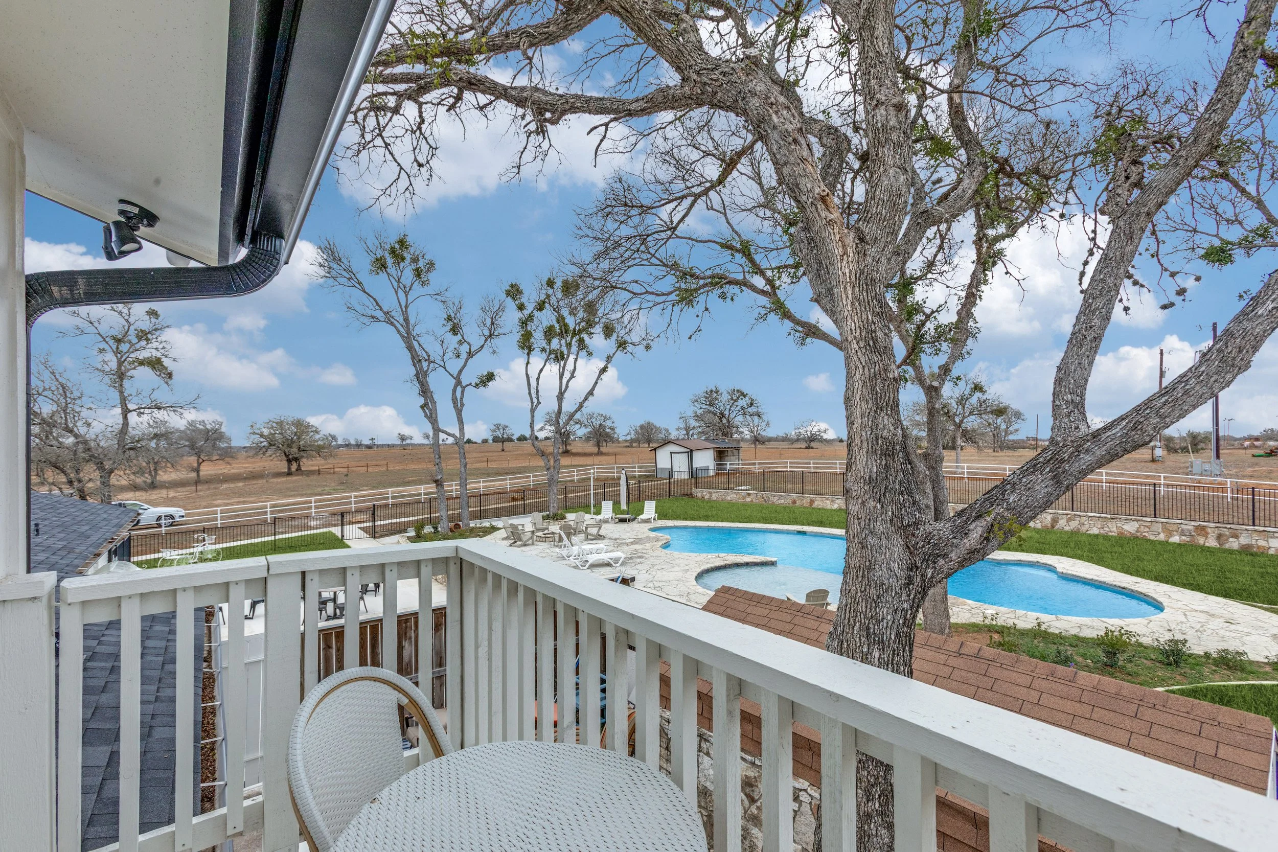 View from a balcony showing a backyard with a swimming pool, patio chairs, large trees, and a rural landscape beyond.