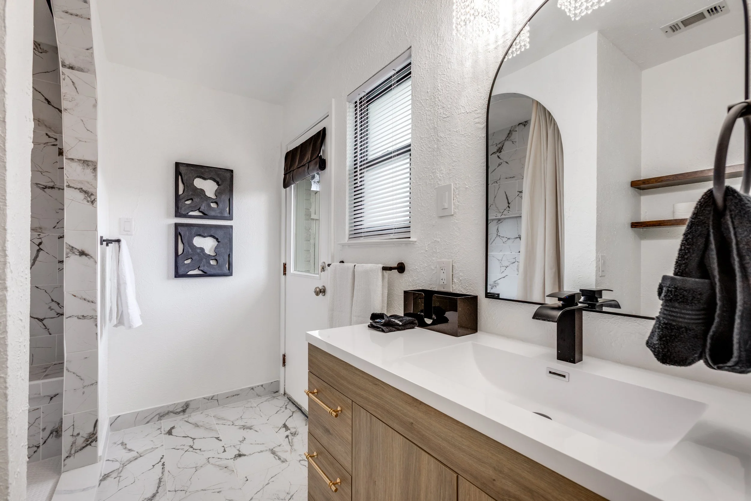 Bathroom with a vanity mirror, marble floor, wooden vanity with gold handles, black faucet, and a black soap dispenser; white walls, a window with blinds, and decorative wall art.