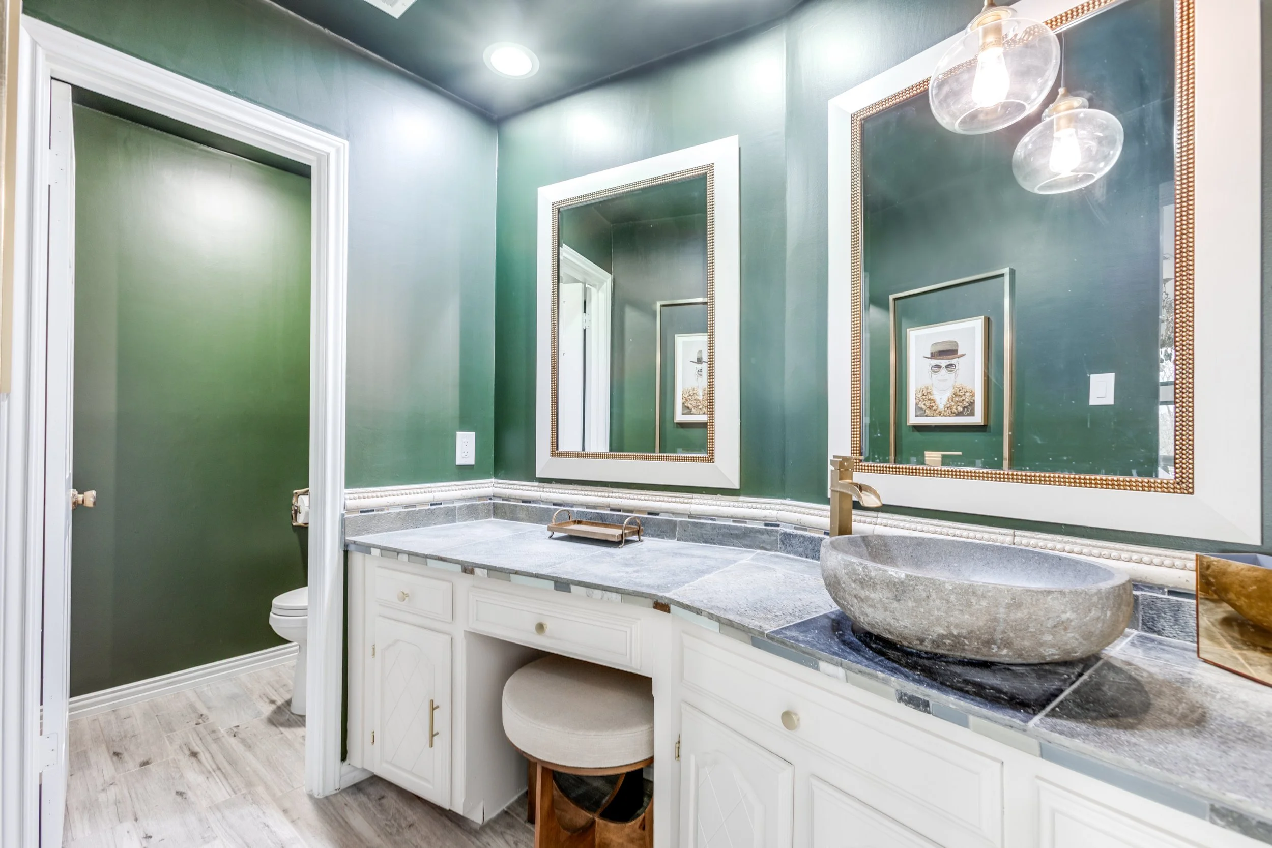 Bathroom with green walls, white cabinetry, a large round stone vessel sink, and framed artwork of a person wearing sunglasses and a hat. There are two glass pendant lights and a large mirror.