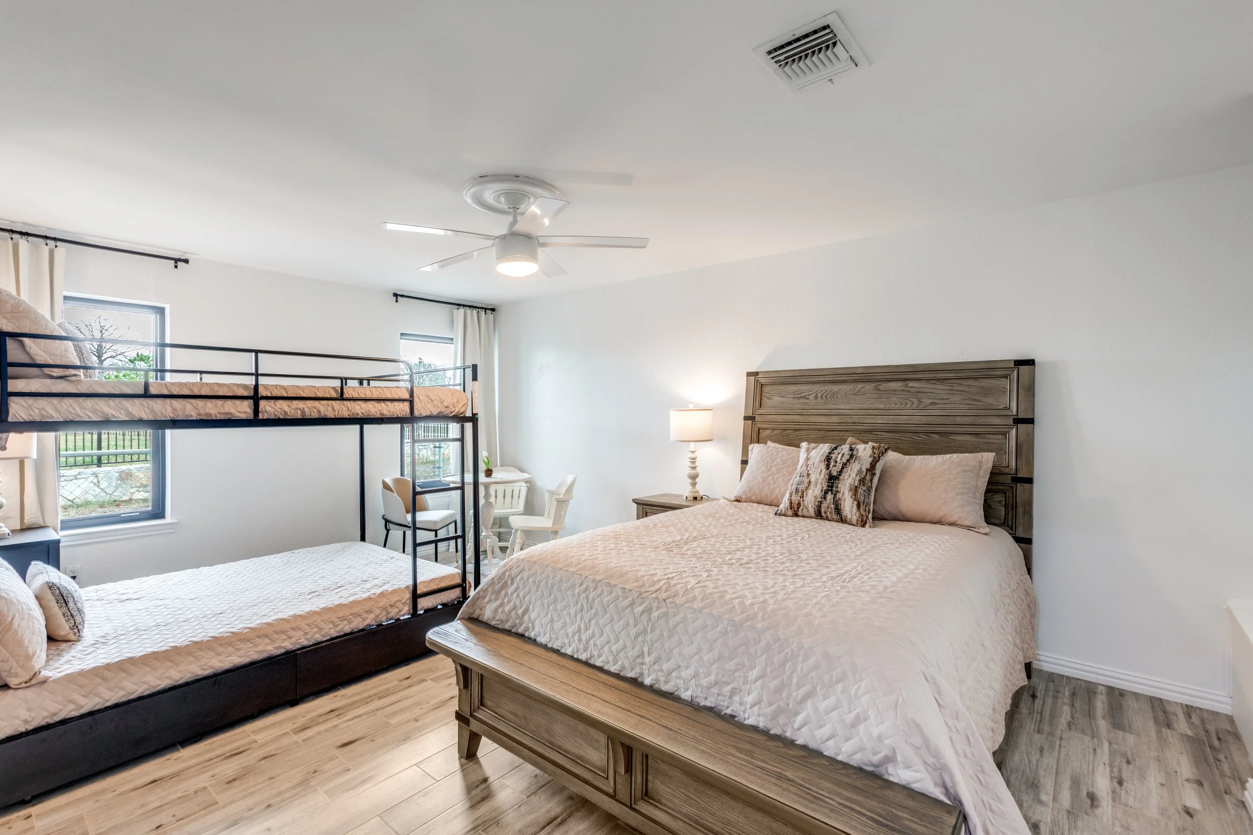 A bedroom with two beds, one with a wooden headboard and the other a metal bunk bed. The room has windows with curtains, a nightstand with a lamp, and a small table with chairs. The ceiling has a fan and an air vent.