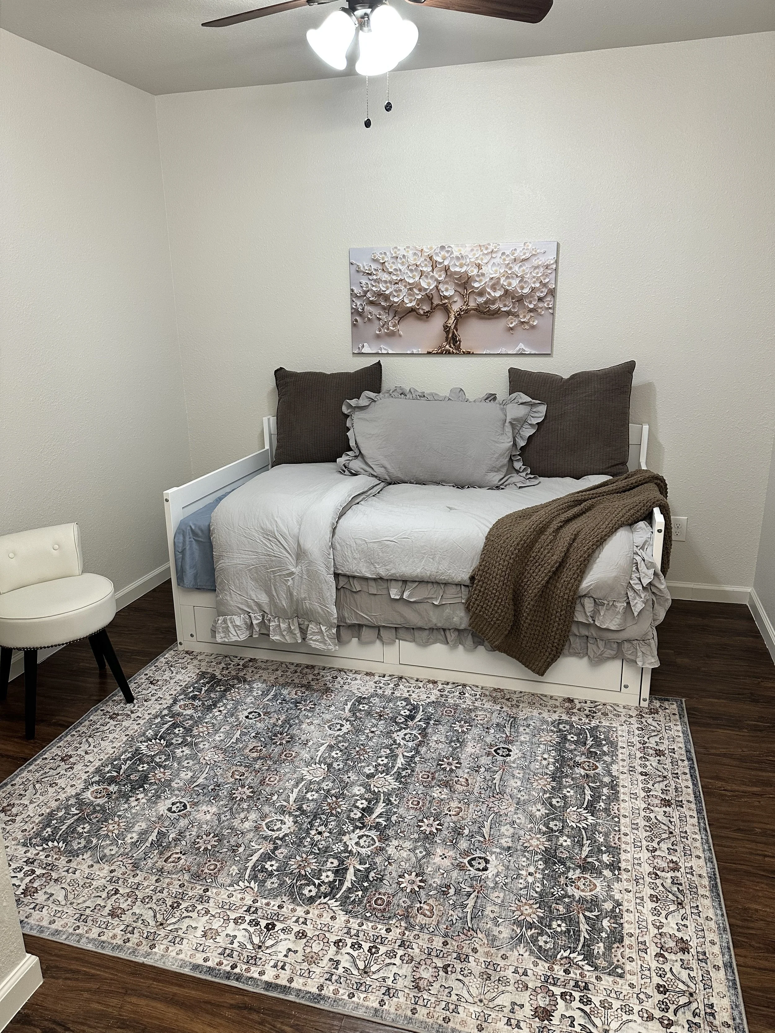 A bedroom with a white bed and ruffled bedding, two large brown pillows, and a gray decorative pillow with ruffled edges. A brown knitted blanket is draped over the foot of the bed. There is a patterned area rug with floral designs in front of the be