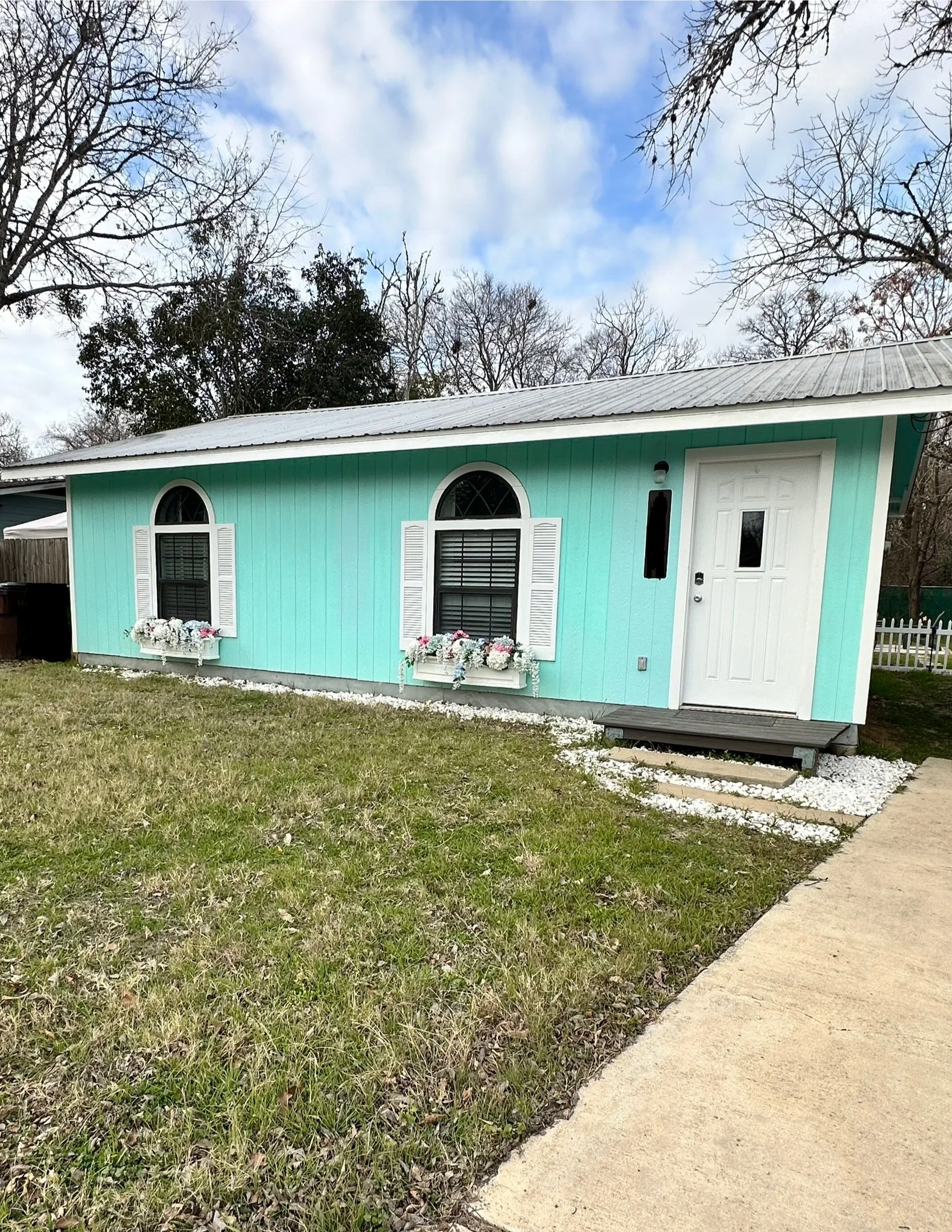 A small turquoise house with white trim, white shutters on the windows, and a white front door. There is a flower box beneath the windows filled with white and pink flowers. The house is surrounded by a grassy lawn and a concrete sidewalk, with leafl