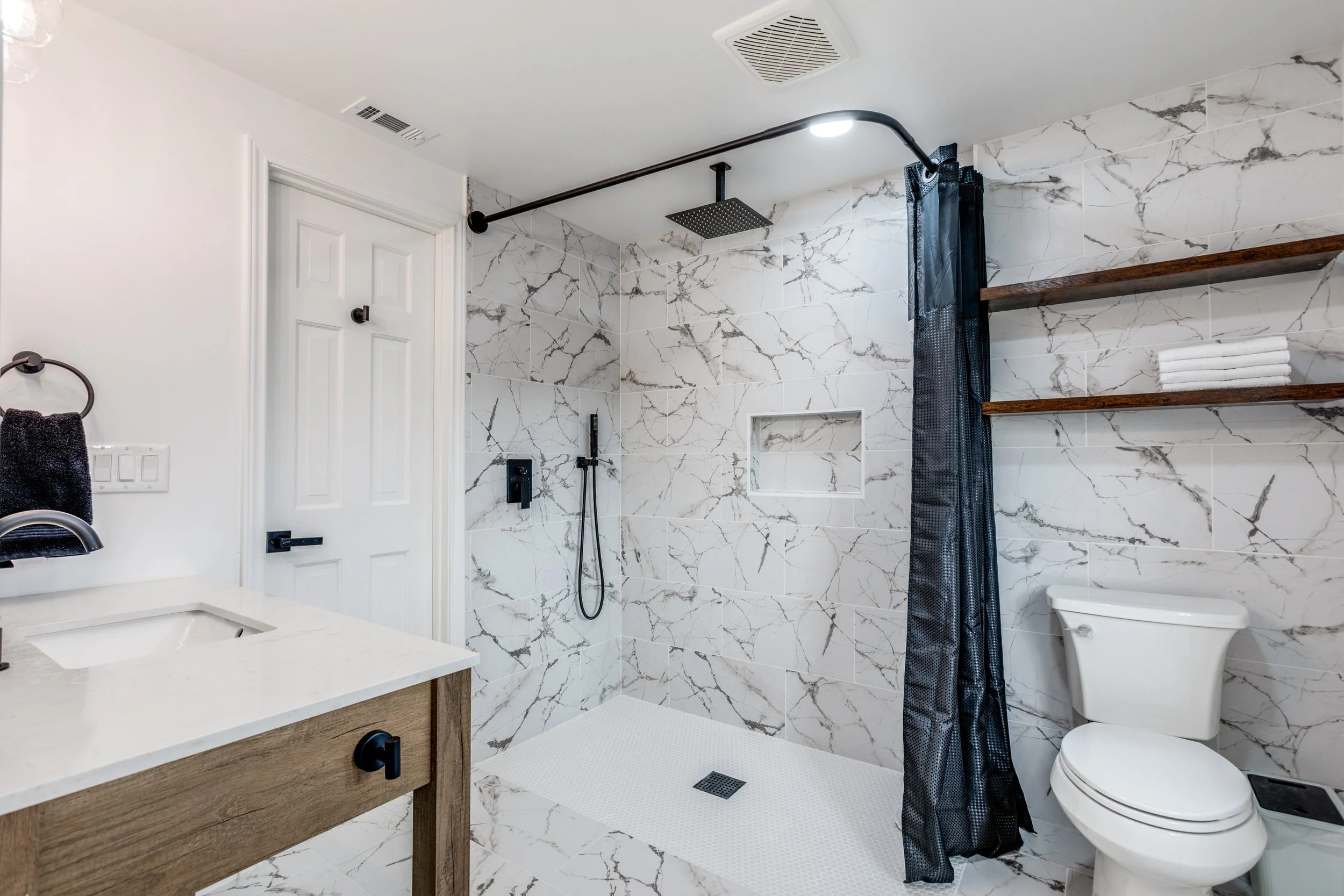 Modern bathroom with white marble walls, a walk-in shower with black fixtures and a rainfall shower head, a toilet, a wooden vanity with a white countertop, black hardware, and open wooden shelves with towels.