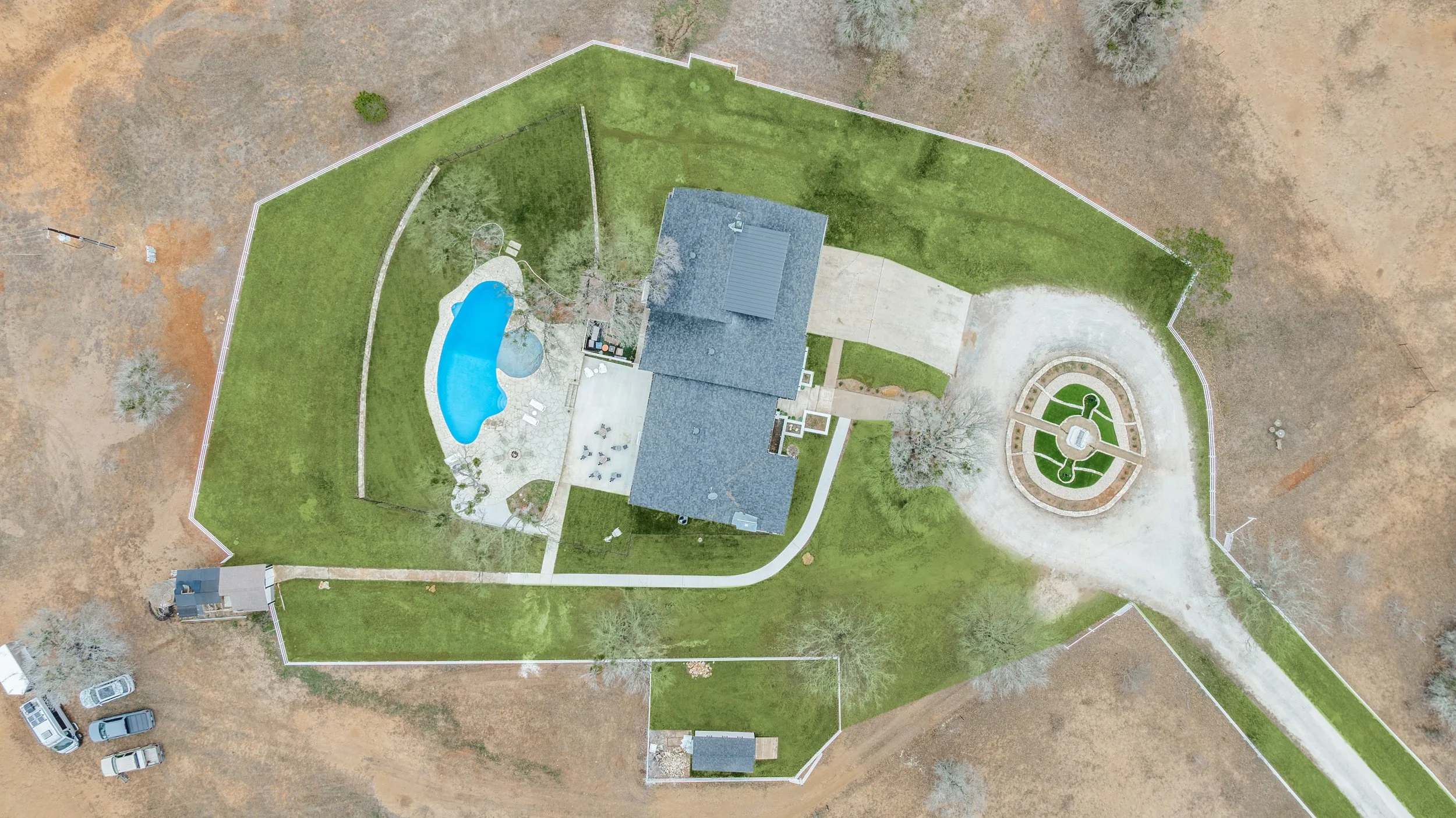 Aerial view of a house with a backyard pool, a green lawn, and a circular garden, enclosed by a white fence.
