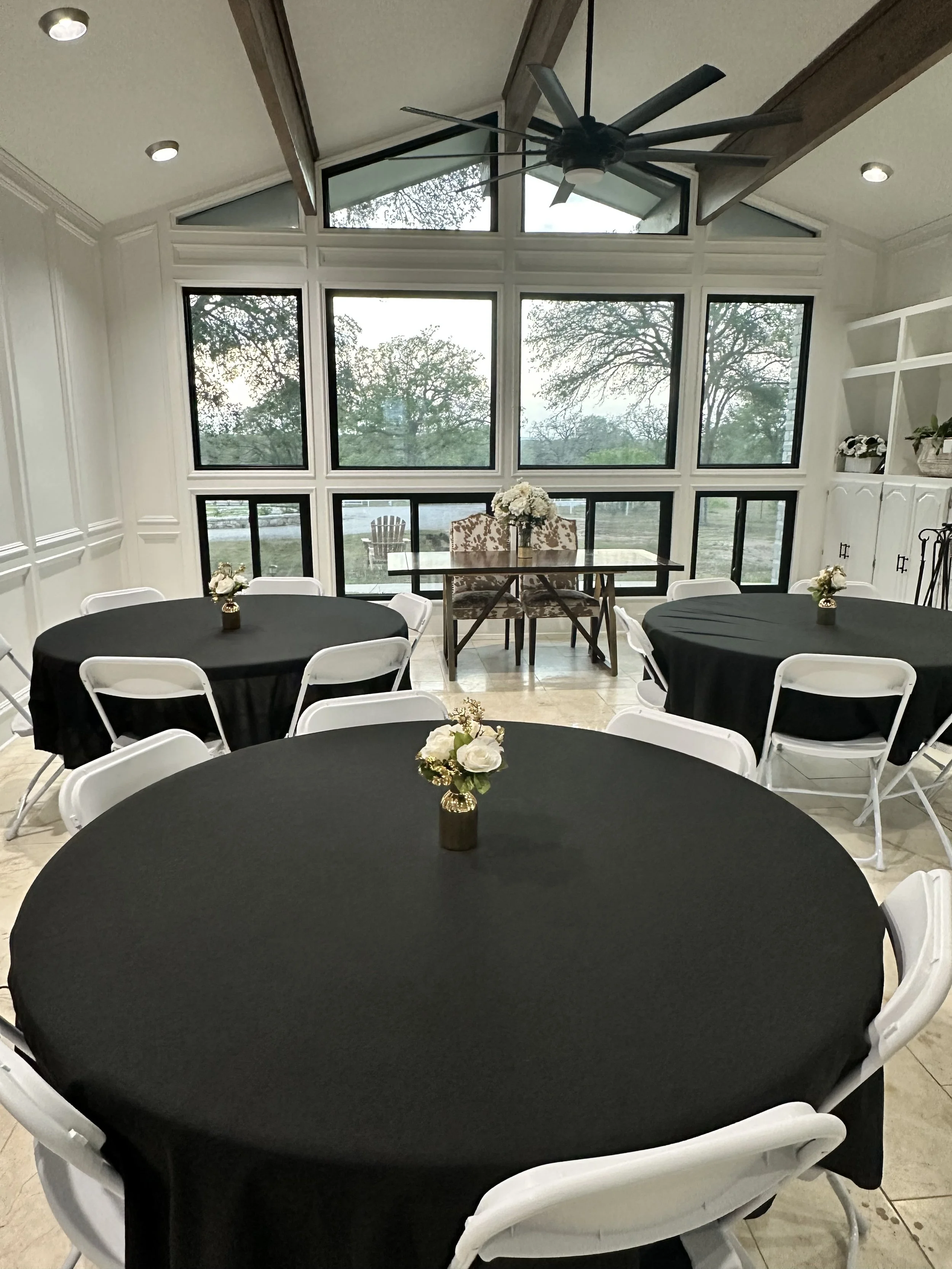 Interior of a room with large windows, round tables covered with black tablecloths, and small floral centerpieces.