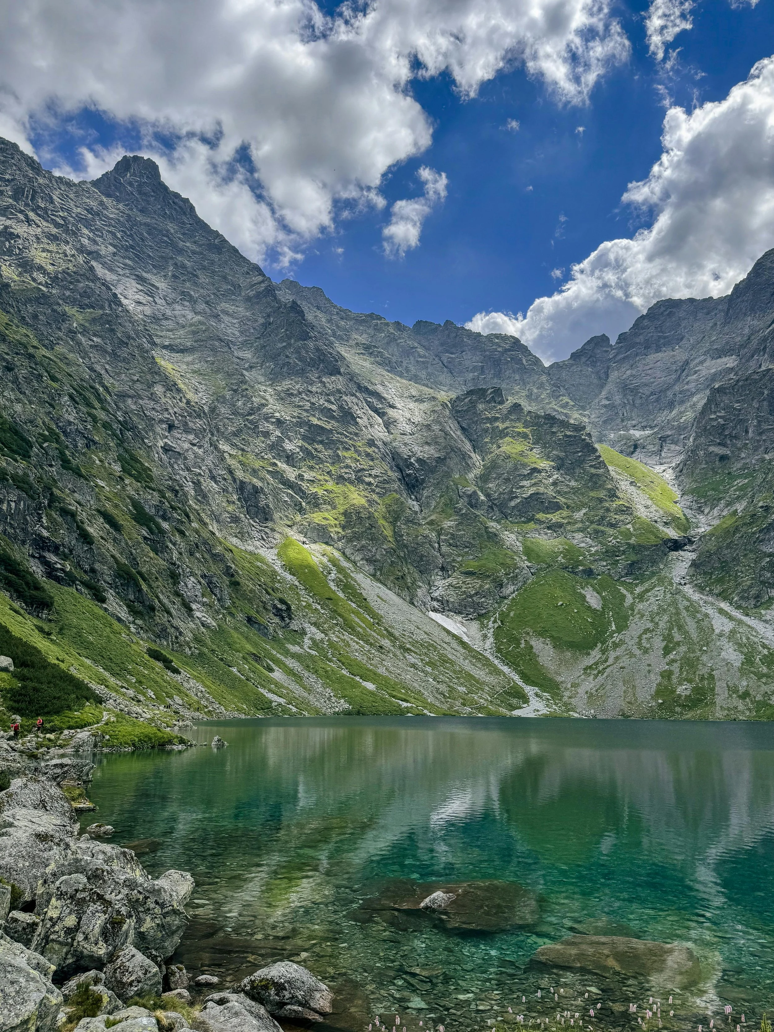 A mountain lake surrounded by tall, rugged mountains with green slopes and patches of loose rock, under a partly cloudy blue sky.