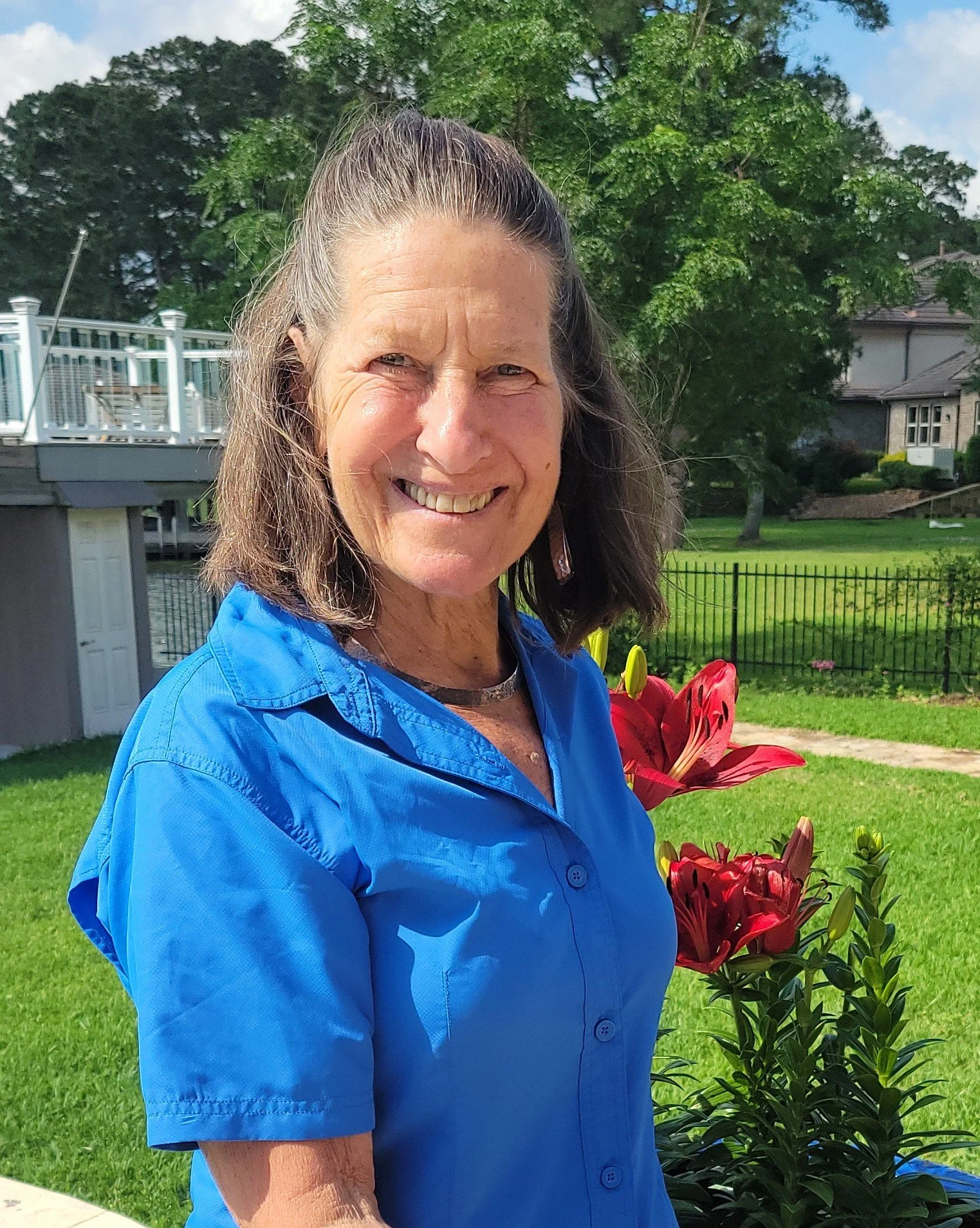A smiling woman wearing a blue shirt holding red flowers outdoors in a backyard with green grass, trees, and a house in the background.